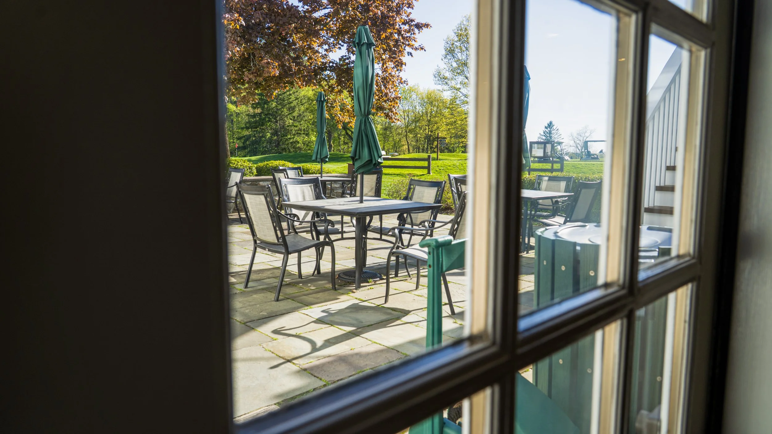 View of an outdoor patio with tables, chairs, and closed umbrellas seen through a window.