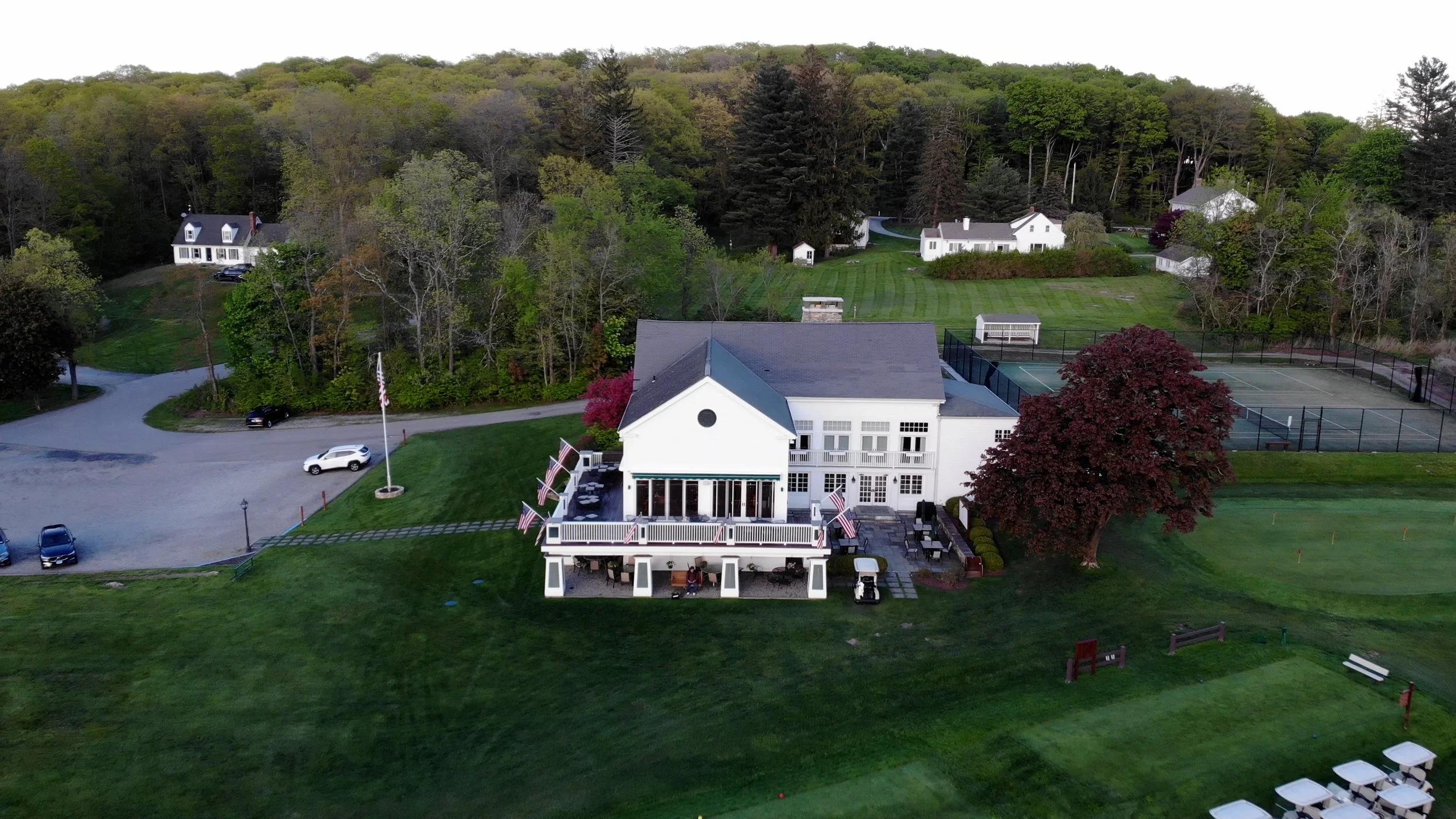 An aerial view of a large white house with a charm-filled porch and an American flag, surrounded by lush green lawns, tennis courts, and trees, with additional houses and forested hills in the background.