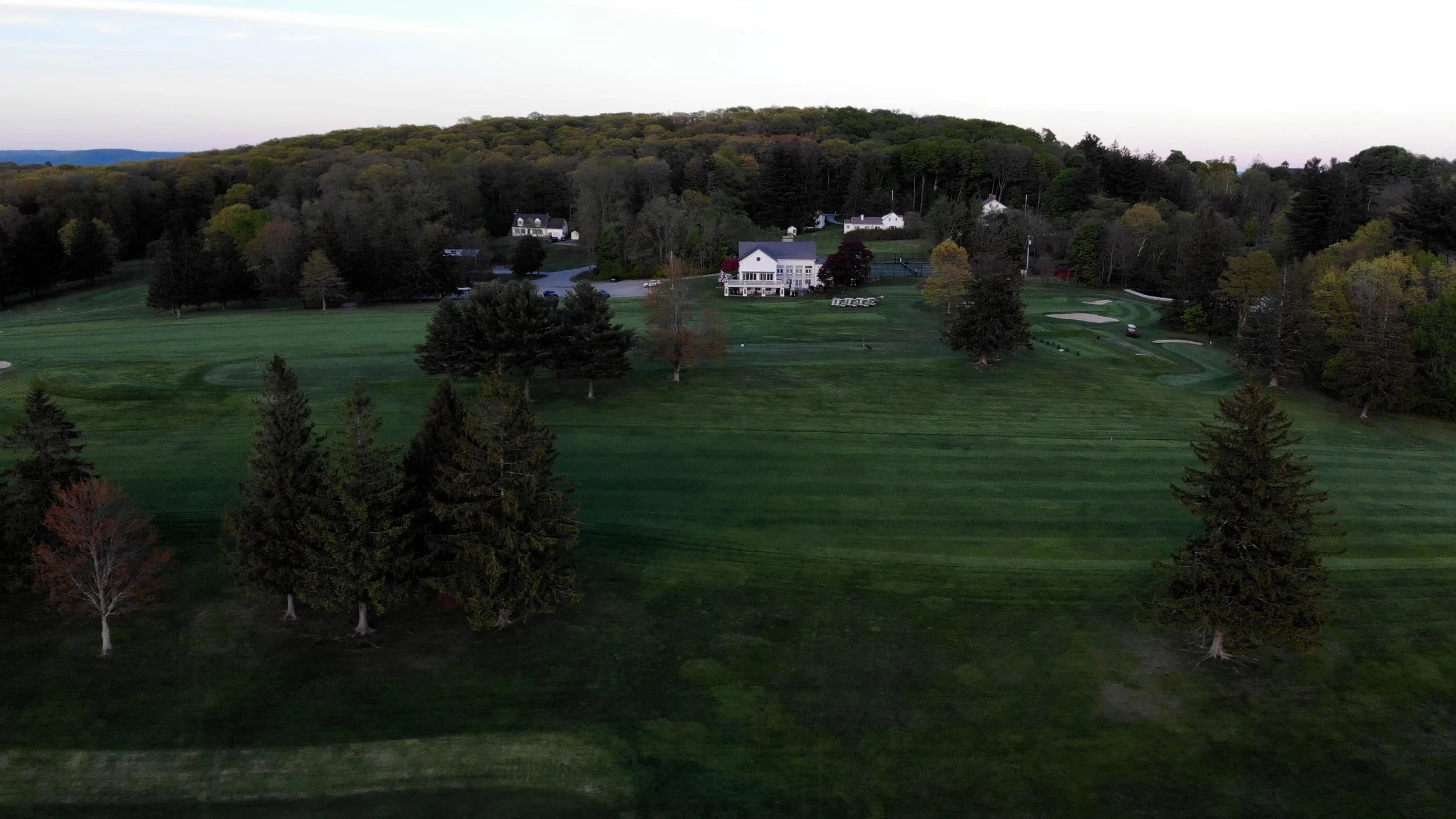 A golf course with lush green grass, surrounded by trees and a few scattered buildings in the background, under a clear sky.