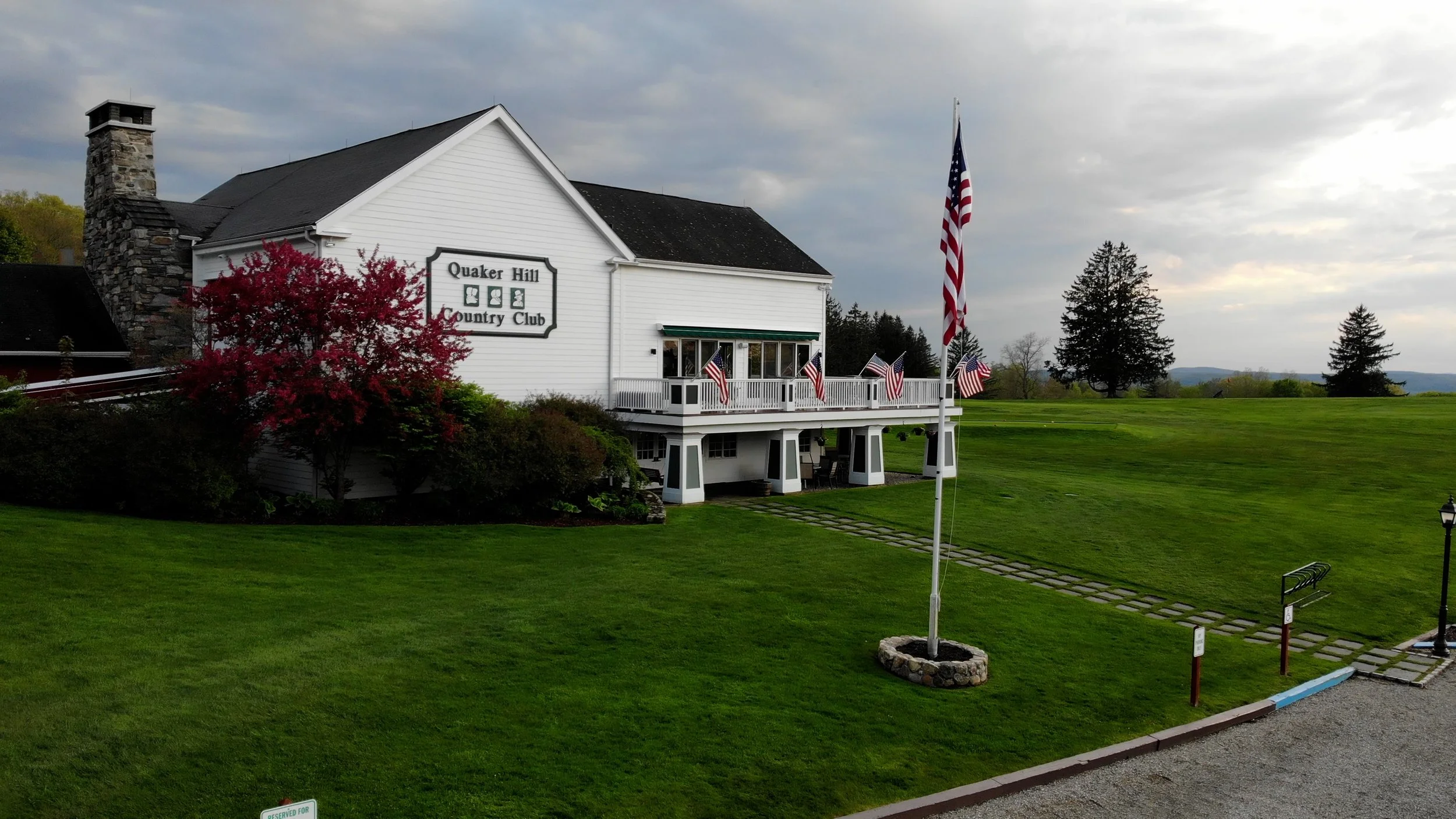 White building labeled 'Quaker Hill Country Club' with green trim, surrounded by a well-manicured lawn, American flags, and trees, with a cloudy sky overhead.