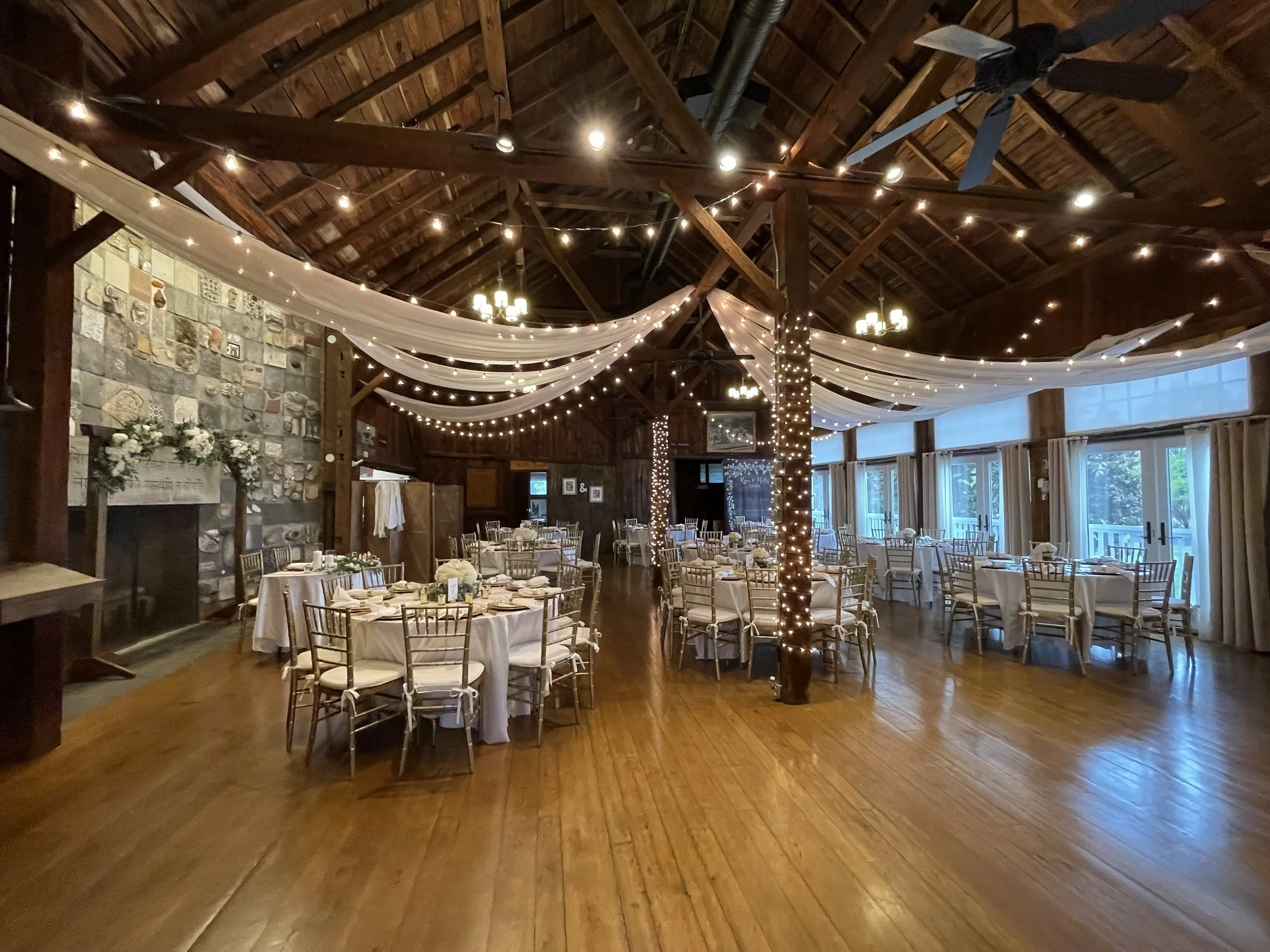 A decorated event space with round tables covered in white tablecloths, gold chairs, and floral centerpieces. The ceiling features draped white fabric and string lights, with wooden beams and a stone fireplace on one wall.