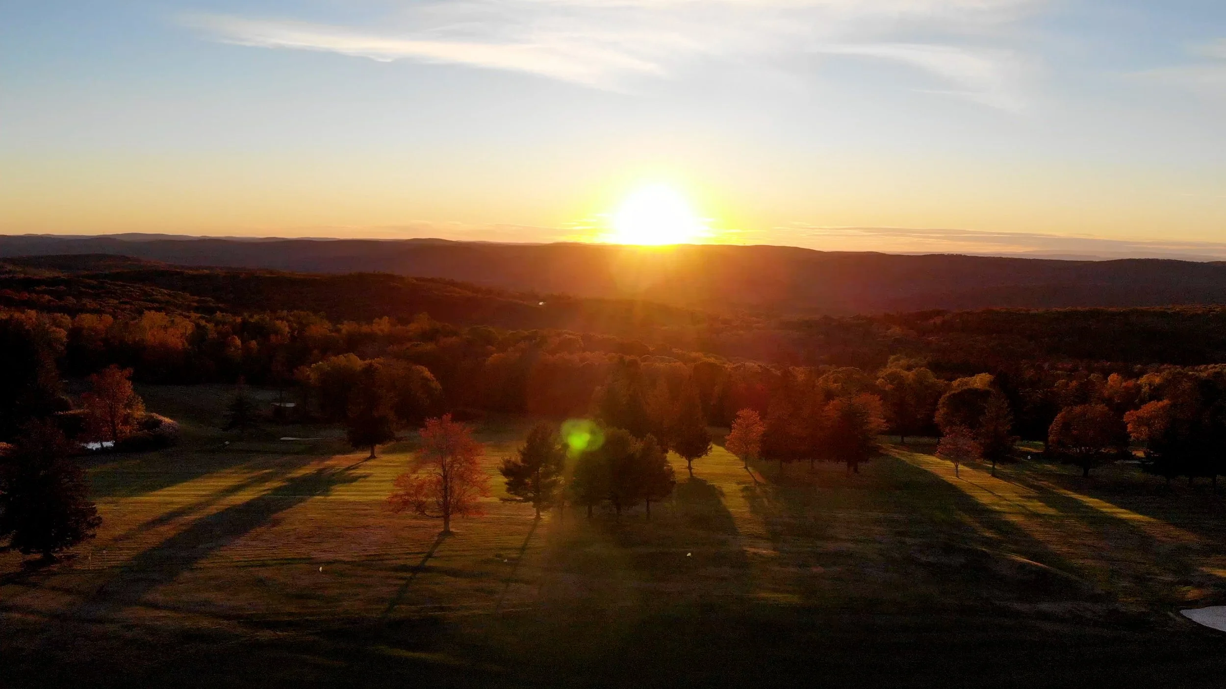 A scenic landscape during sunset showing rolling hills, trees casting long shadows, and a sky with scattered clouds.