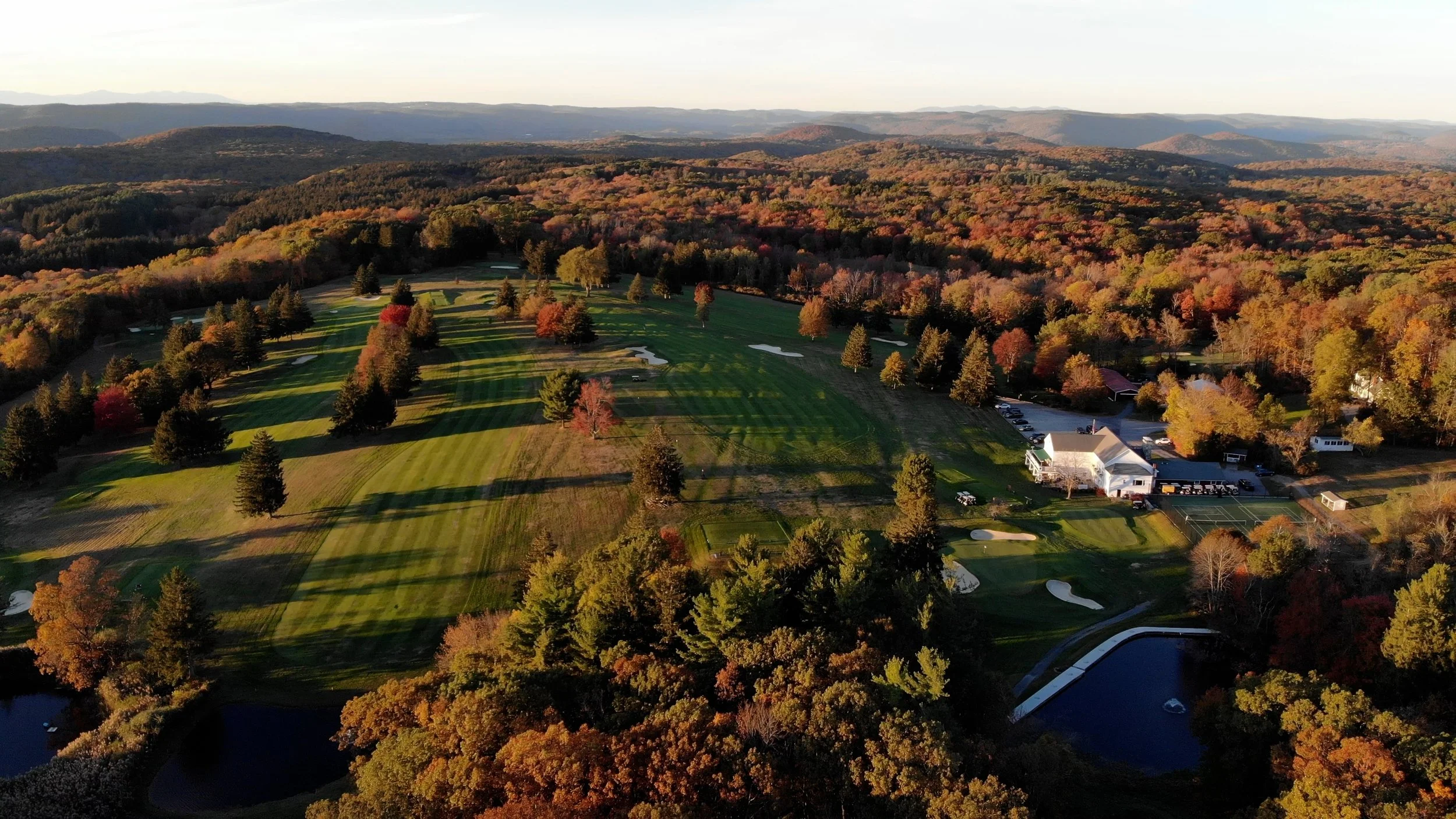 Aerial view of a golf course surrounded by trees with colorful autumn foliage, mountains in the background, and a few buildings and parked cars near the golf course.