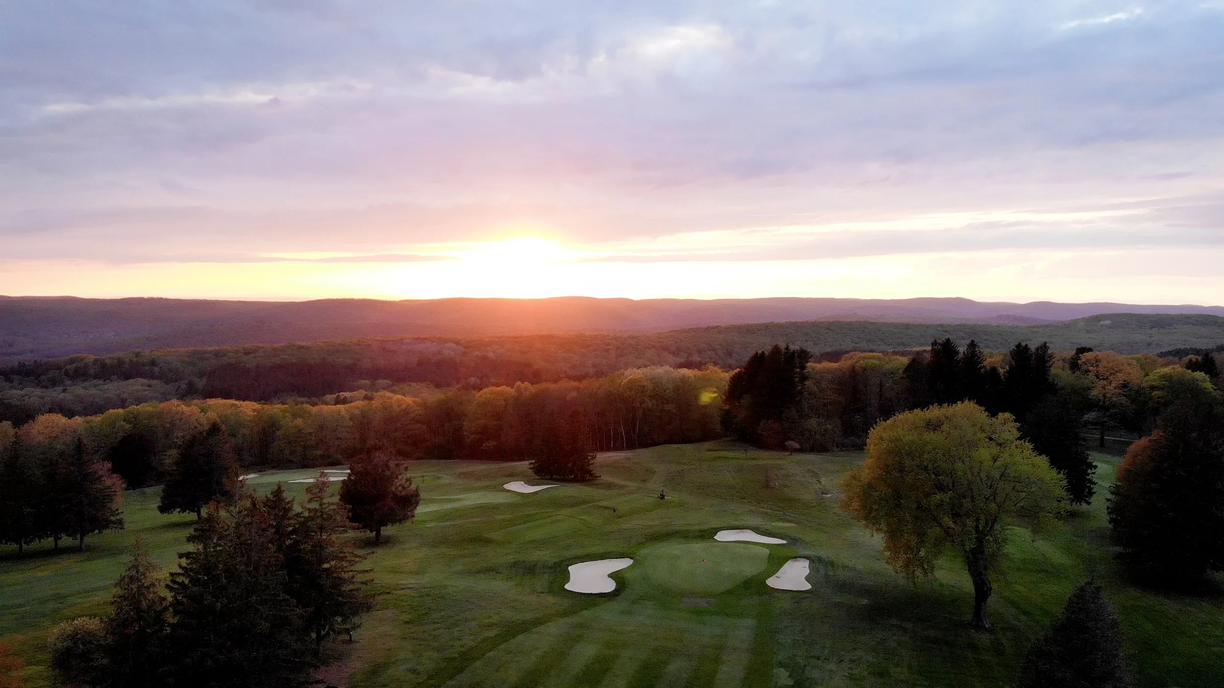 A scenic view of a golf course at sunset with green fairways, sand bunkers, surrounded by trees and rolling hills in the distance under a cloudy sky.