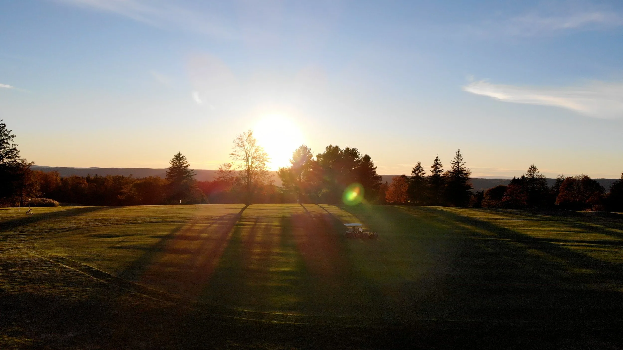 A golf course at sunset with trees casting long shadows and a golf cart on the grass.