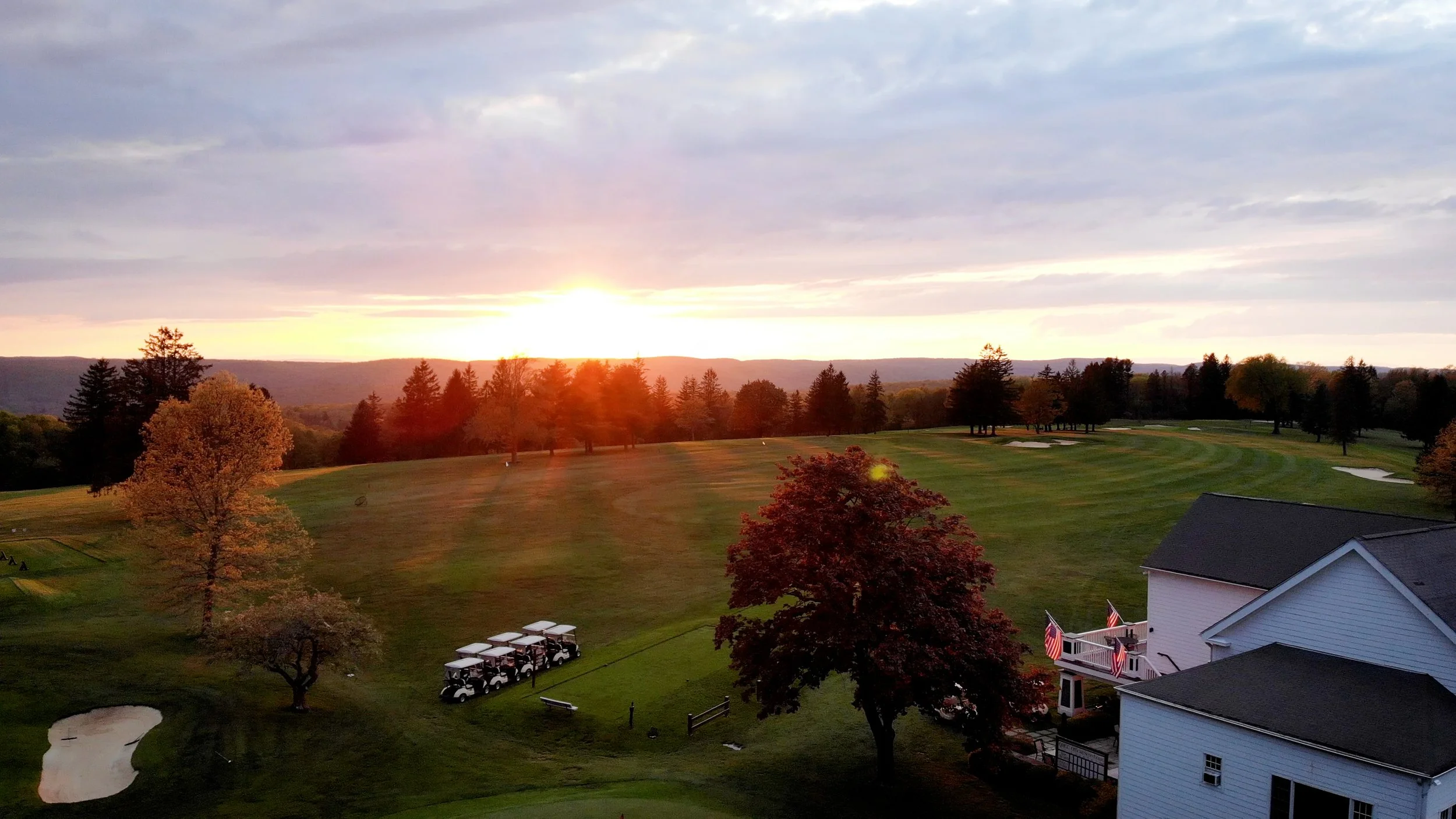 Sunset over a golf course with trees, sand traps, and golf carts, near a house with American flags.