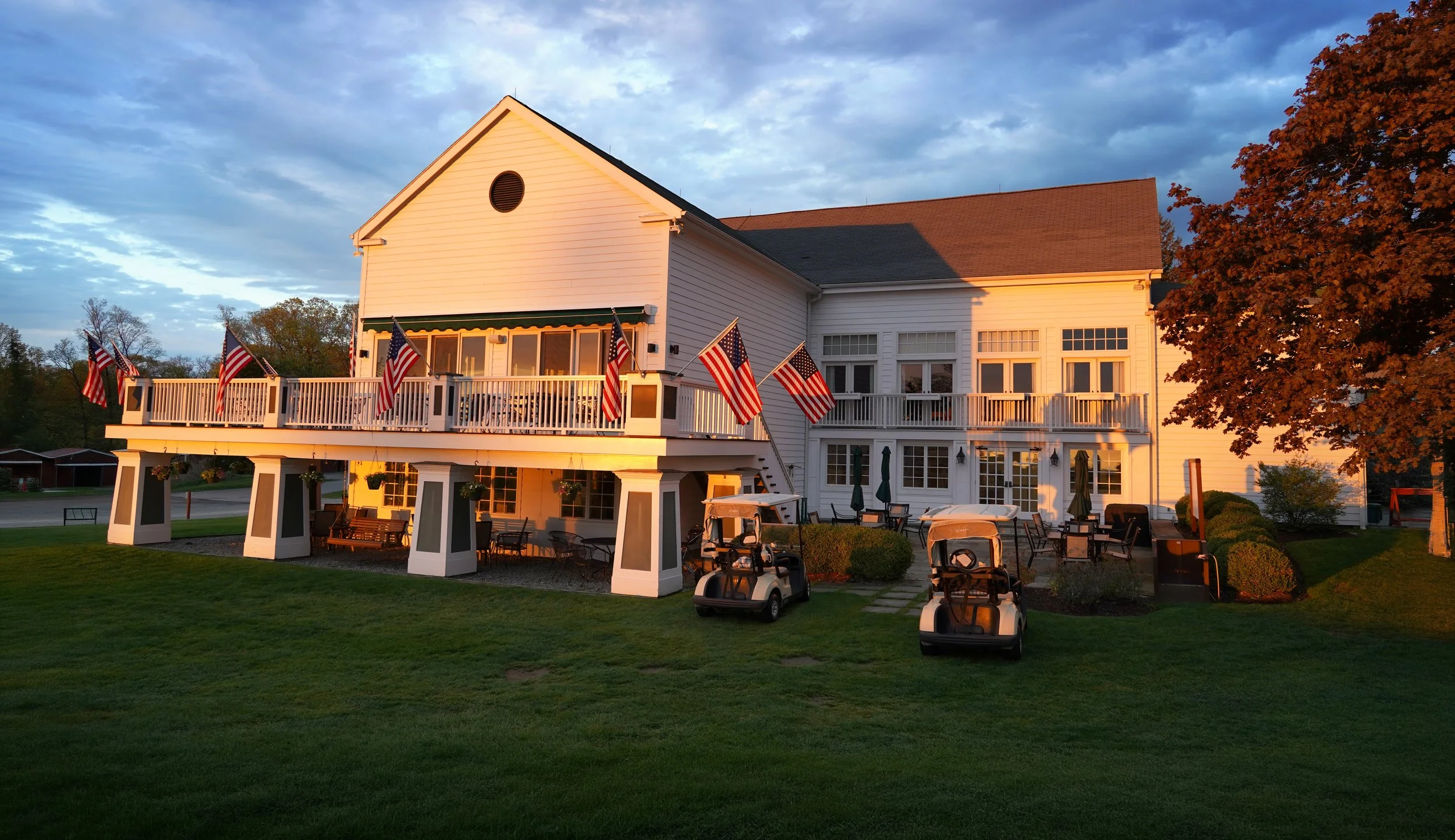 A large white building with a deck decorated with American flags. The area has outdoor furniture and golf carts, with trees and a cloudy sky in the background during sunset.