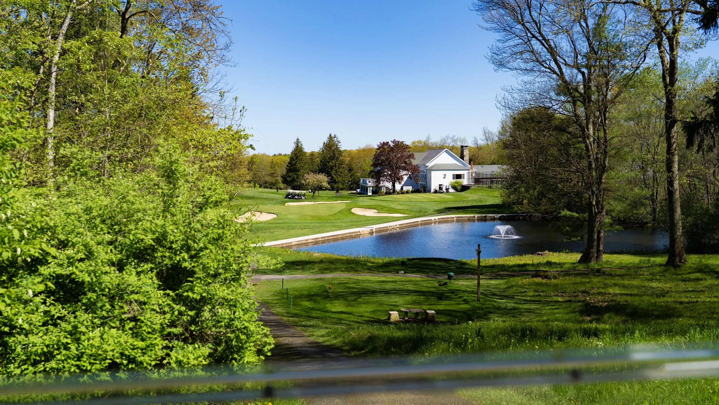A golf course with a pond, sand bunkers, and a white clubhouse surrounded by trees and greenery on a sunny day.