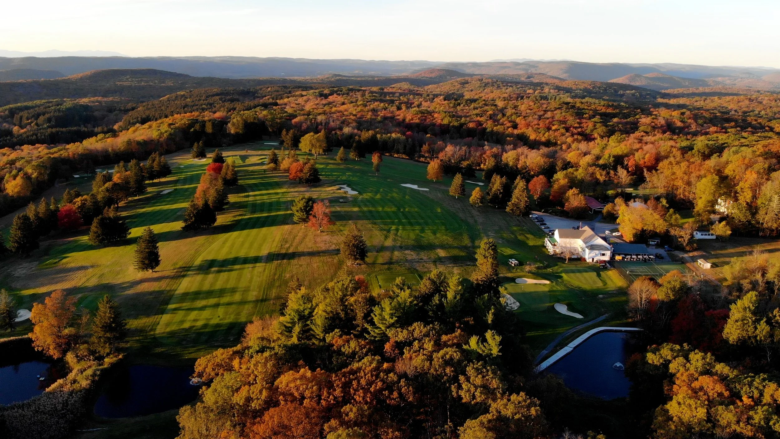 Aerial view of a golf course surrounded by trees with fall foliage, hills in the background, and a few buildings near the course.