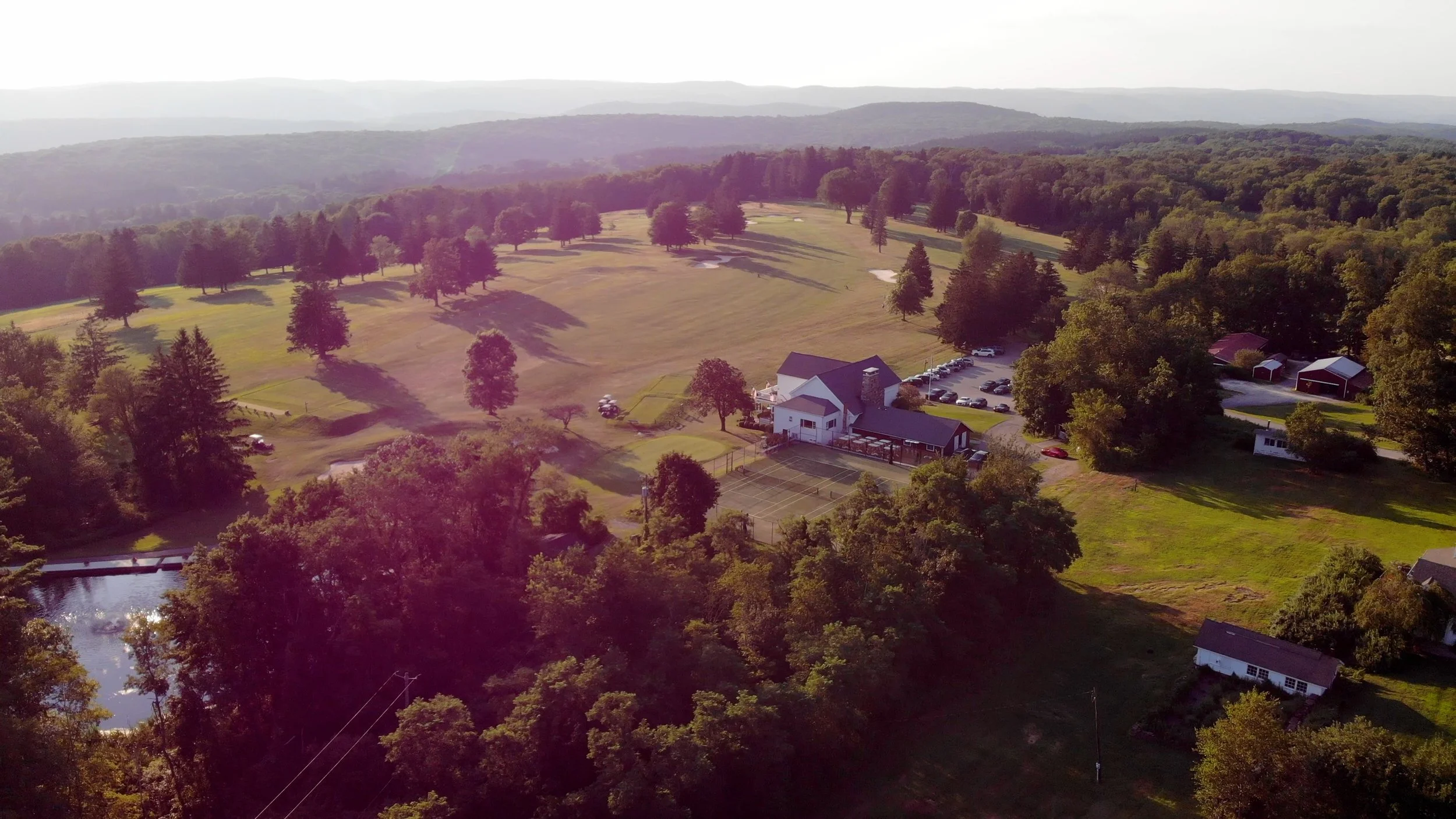 An aerial view of a golf course surrounded by trees, buildings, and a small pond with a bridge, with mountains in the background.