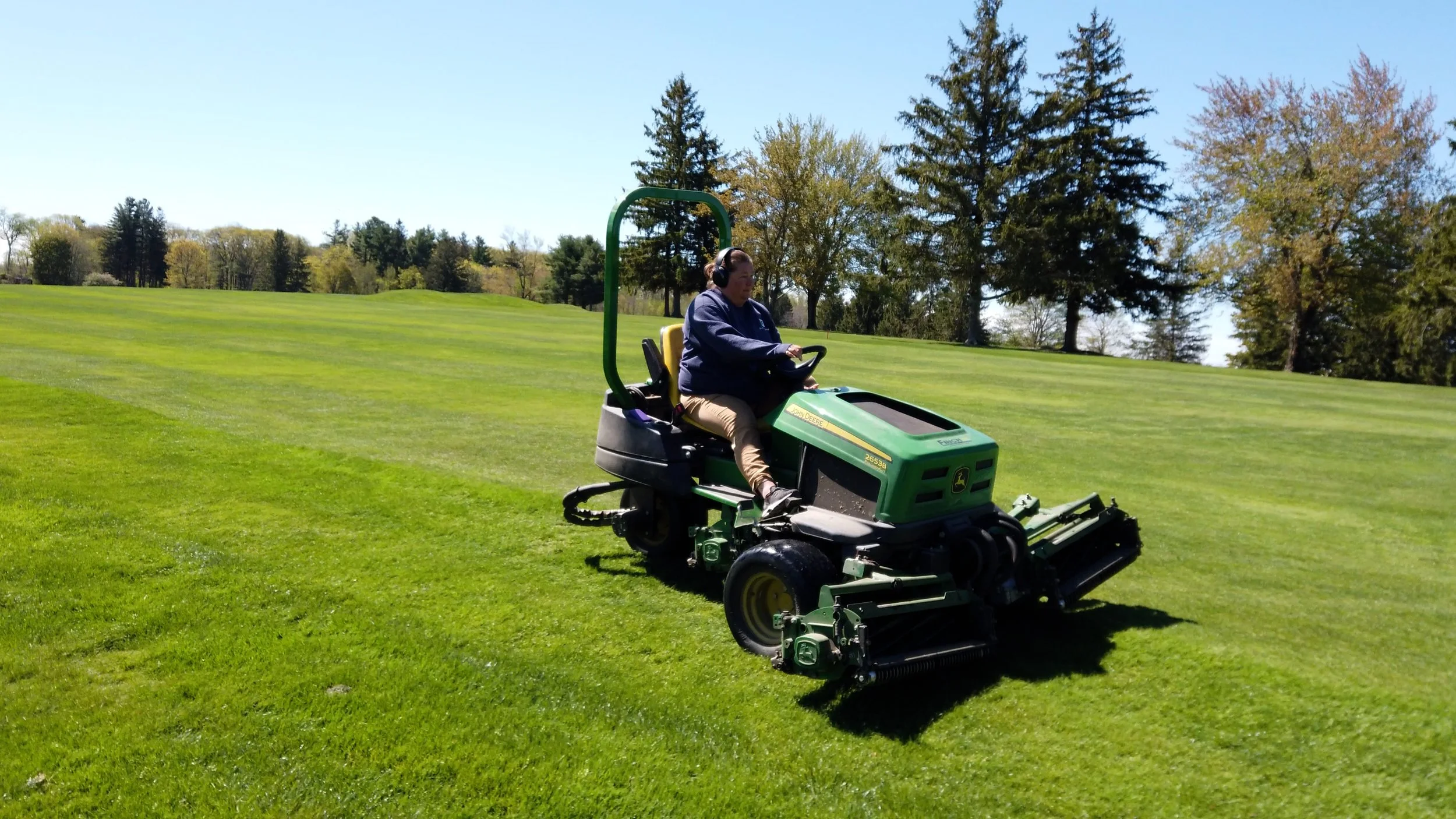 A person operating a green John Deere lawn mower on a well-manicured grassy field with trees in the background and a clear blue sky.