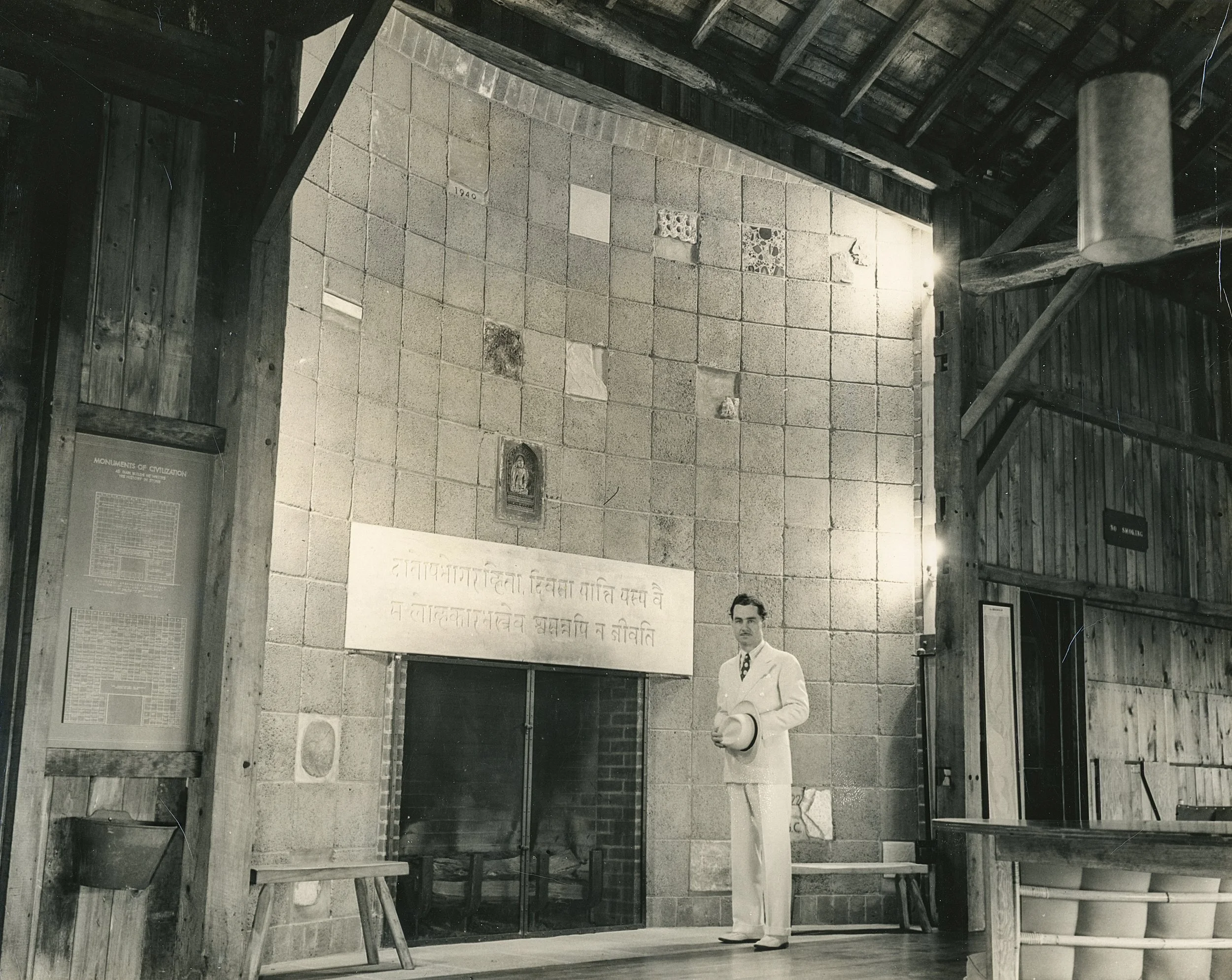 A black-and-white photograph of an indoor space with a man standing near a corner. The space has a brick wall with plaques and inscriptions, and is surrounded by a rustic wooden structure with a sloped roof. A sign says "NO SMOKING."