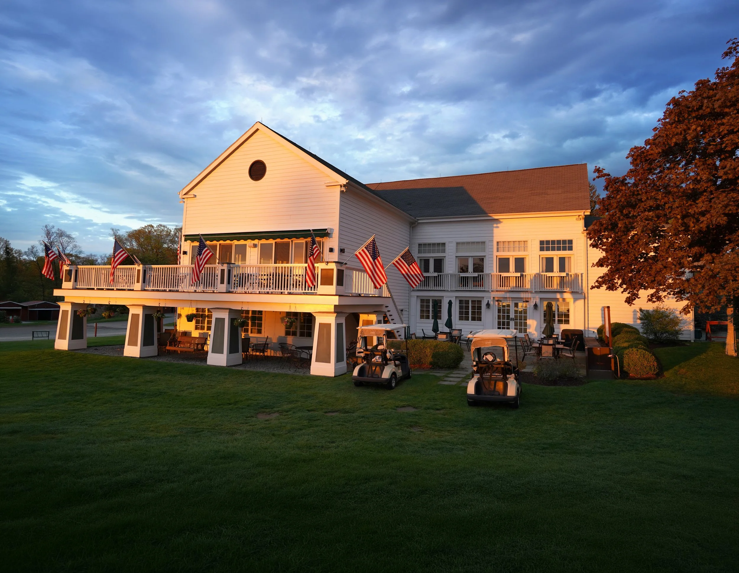 A white house with a large porch decorated with American flags and outdoor furniture, under a sky with clouds and the setting sun.