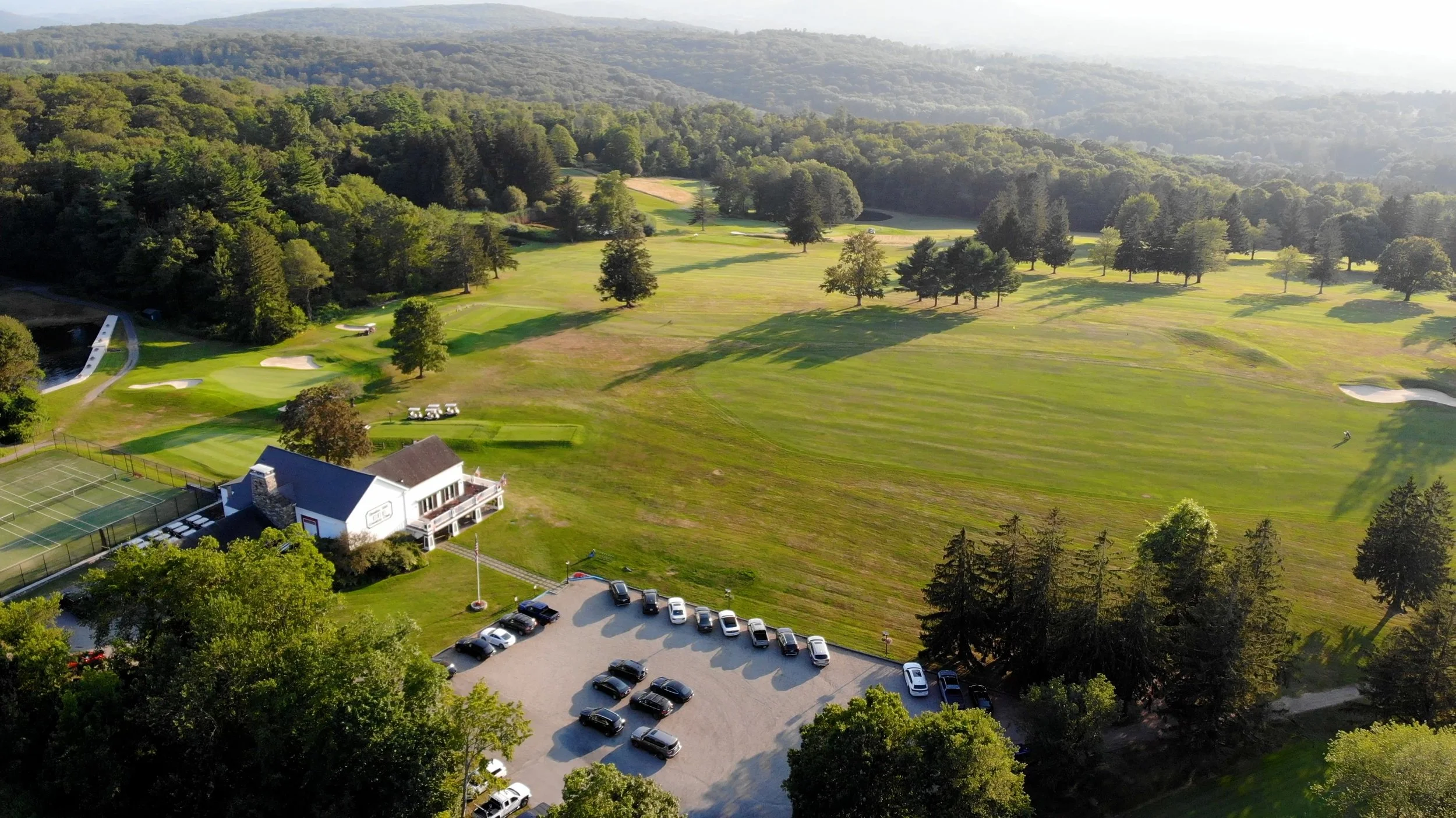 Aerial view of a golf course with several sand traps, trees, and a clubhouse with parking lot filled with cars, surrounded by lush green landscape and forested hills in the background.
