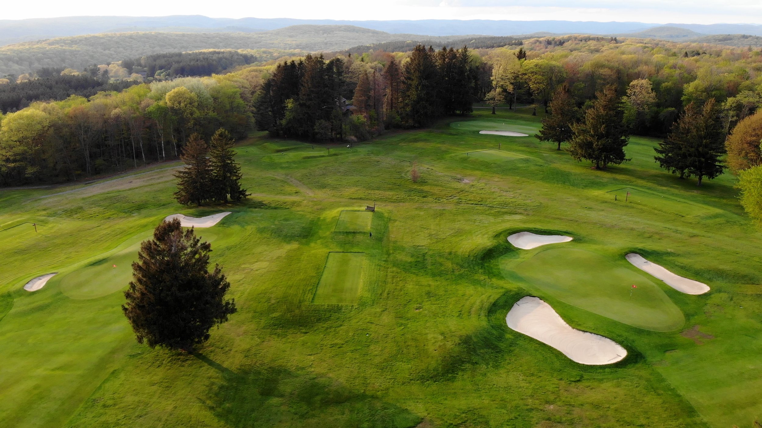 Aerial view of a golf course with green fairways, sand traps, and a wooded area in the background under a cloudy sky.