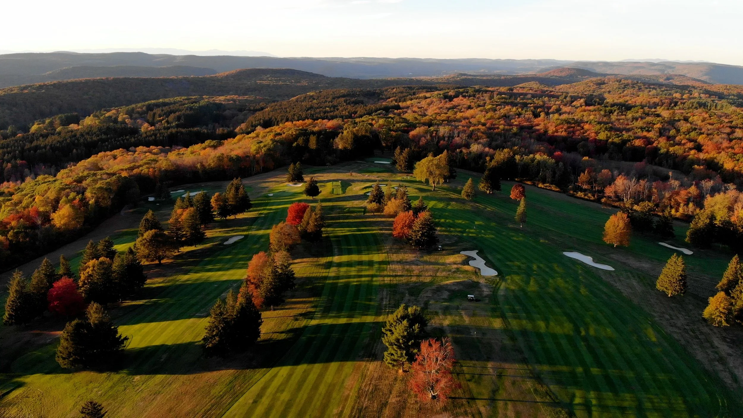 Aerial view of a golf course surrounded by autumn trees, with a mountain range in the distance during sunset.