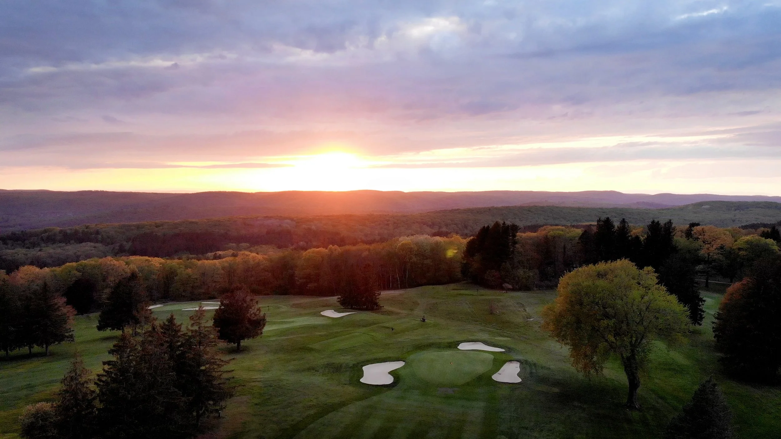 Sunset over a golf course with trees, sand traps, and a green, surrounded by a forested landscape.