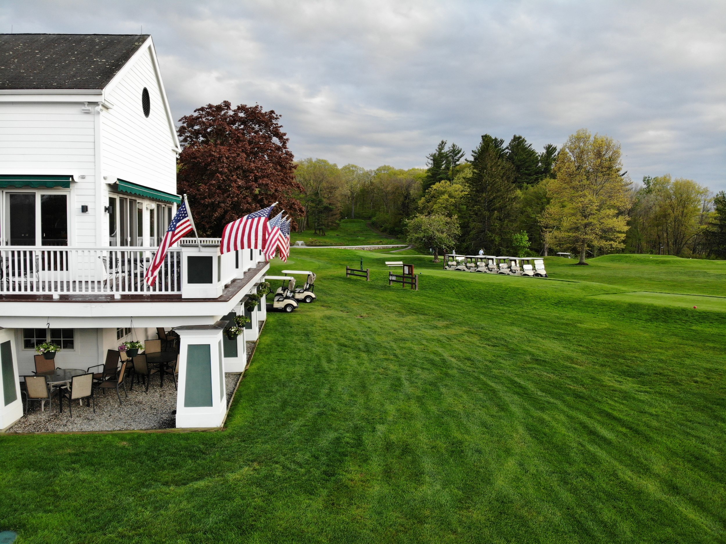 A white building with a deck decorated with American flags, outdoor seating beneath green awnings, a lush green lawn, trees, and a cloudy sky.