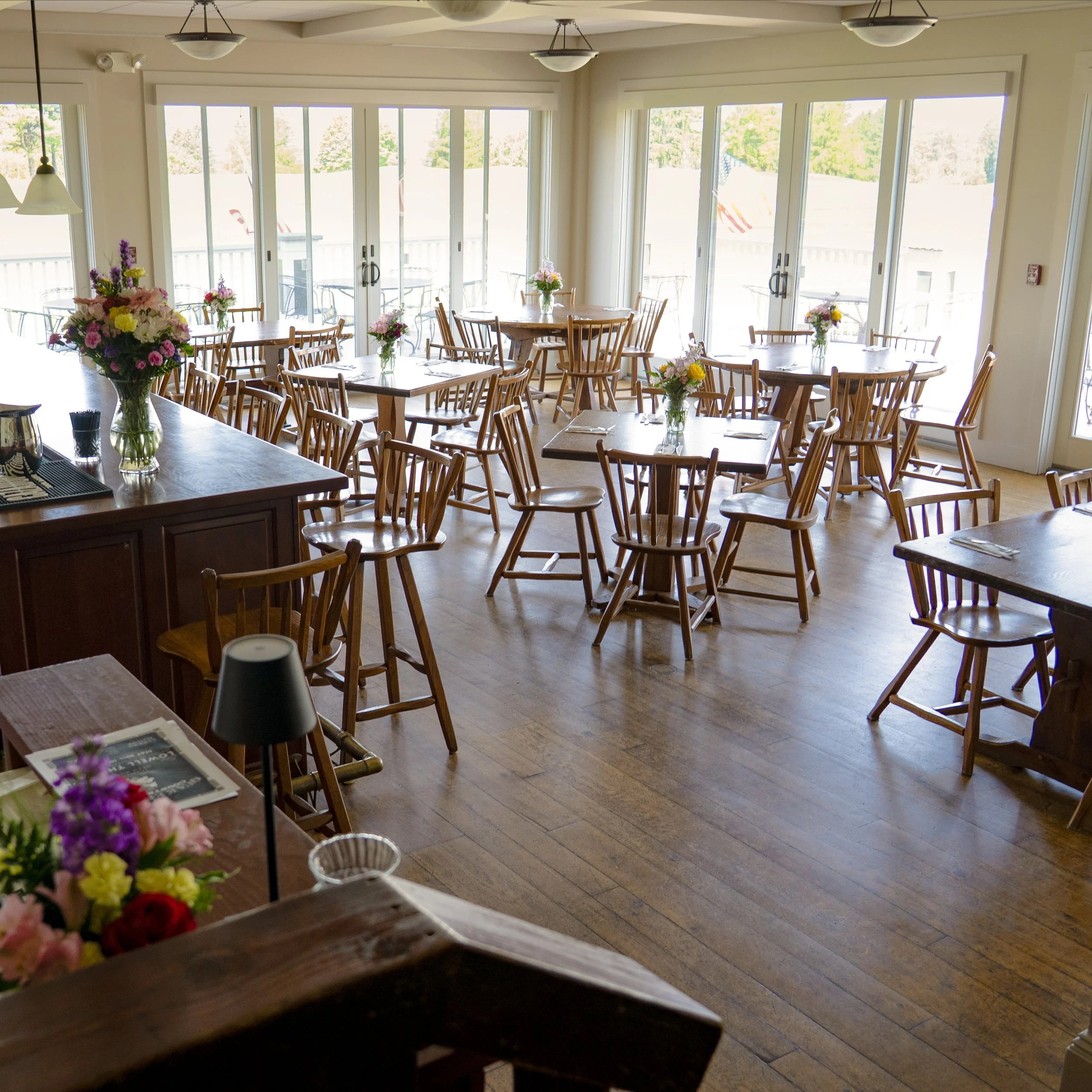 Interior of a bright restaurant or cafe with wooden tables and chairs, decorated with vases of colorful flowers, large windows with a view of trees and outdoor seating.