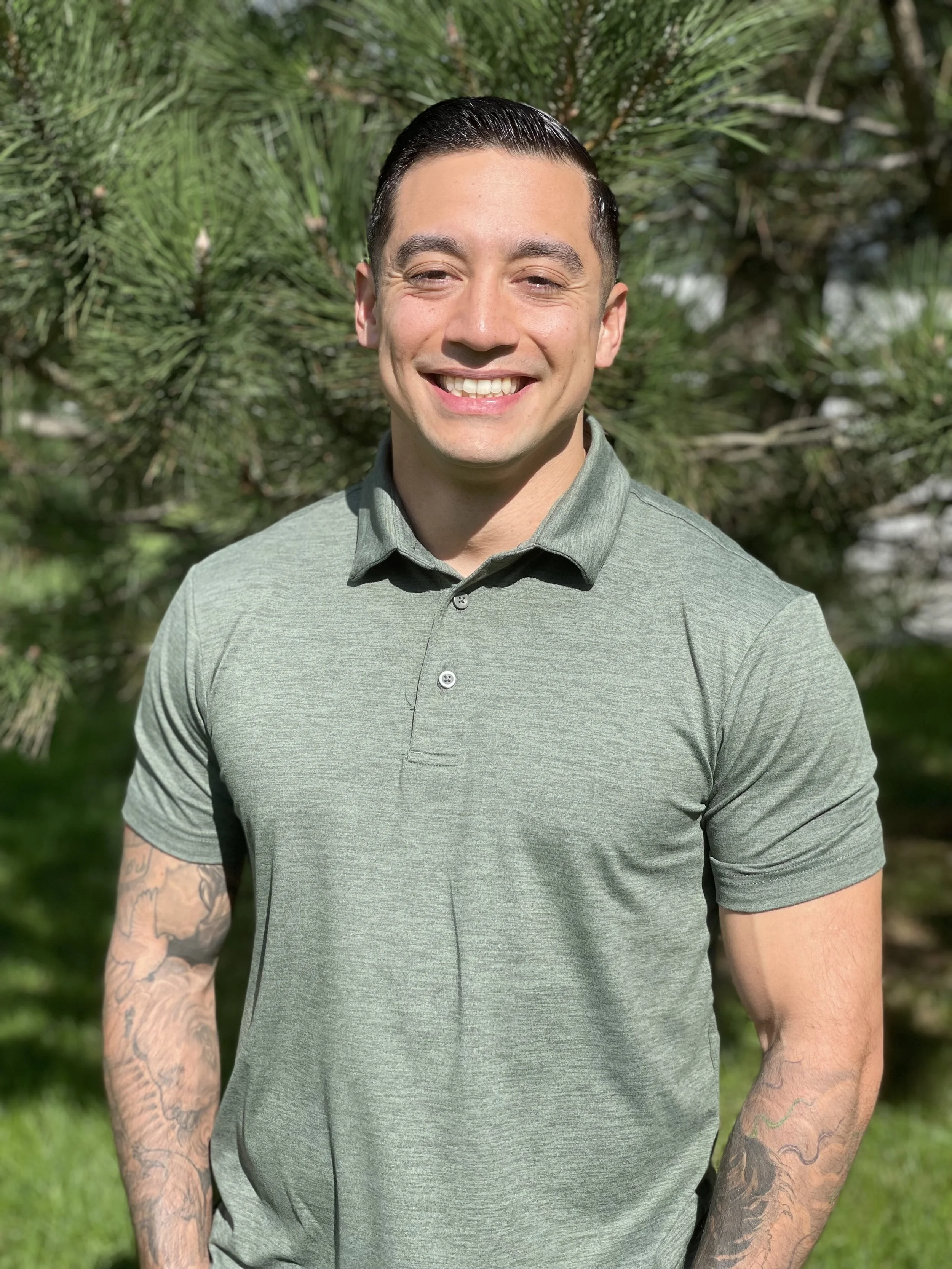 A young man with dark slicked-back hair smiling outdoors in front of a green pine tree, wearing a light green polo shirt with tattoos on his arms.