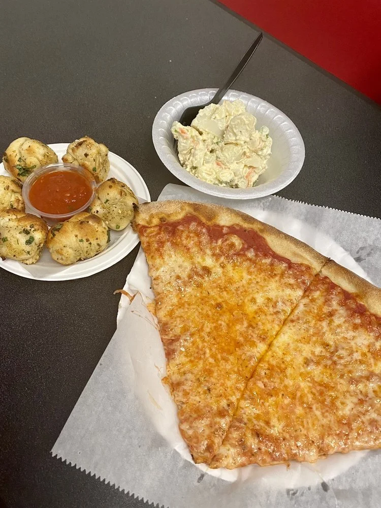 A slice of pepperoni pizza on white paper, a small plate of garlic knots with marinara sauce, and a side of potato salad in a foam bowl with a spoon.