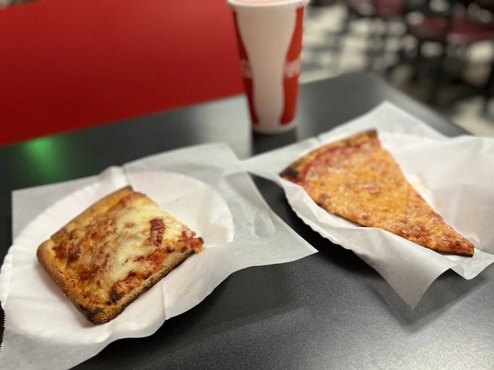 Two slices of pizza on paper in front of a soda cup on a black table.