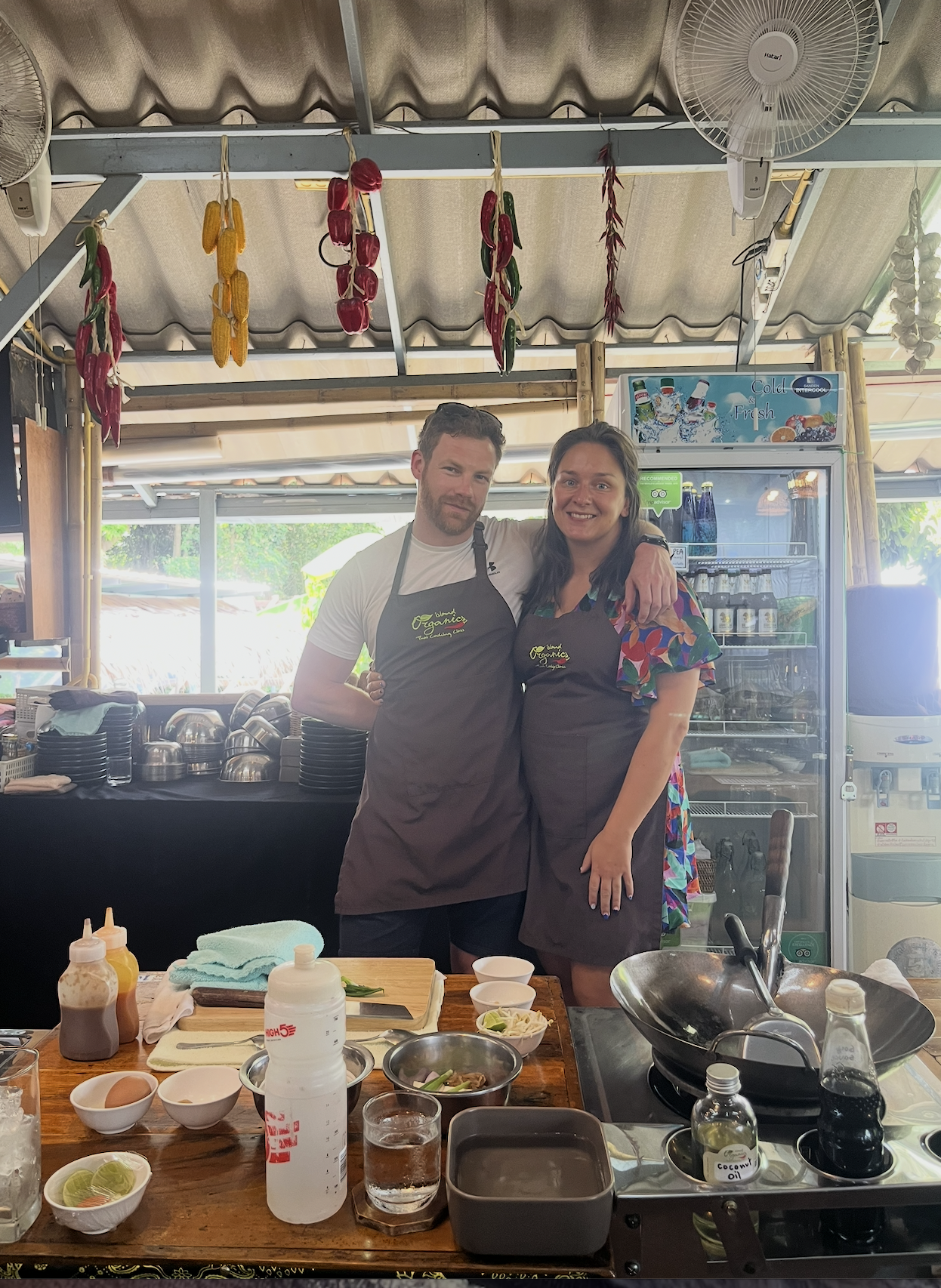 Two people, a man and a woman, standing in a kitchen or cooking area, smiling and wearing aprons. The area has various cooking ingredients and utensils on the table in front of them, with dried chili peppers and corn hanging from the ceiling.