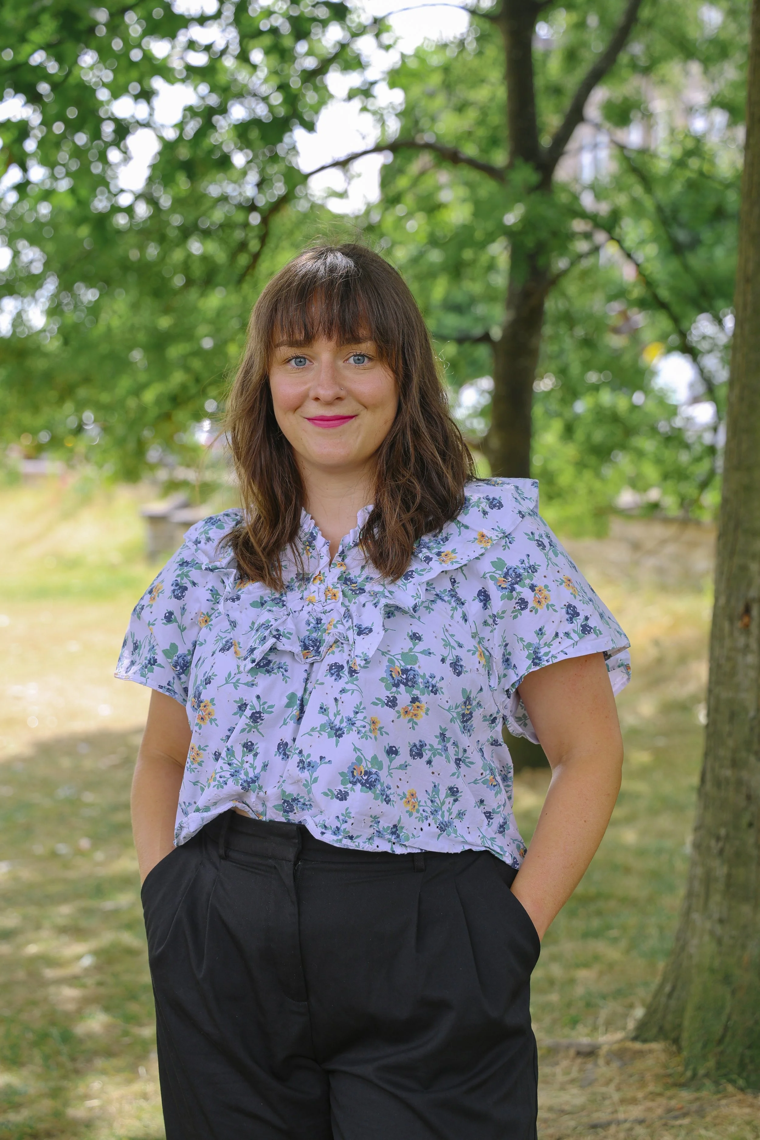 A woman with brown hair and blue eyes, wearing a floral blouse and black pants, standing outdoors in a park with green trees in the background.