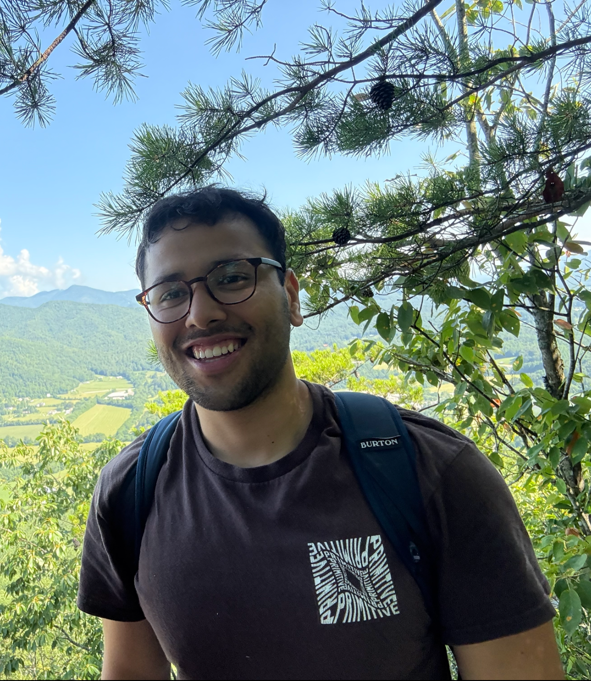 A smiling man with glasses and a backpack standing outdoors in a lush, green landscape with mountains in the background, under a tree with pine cones.