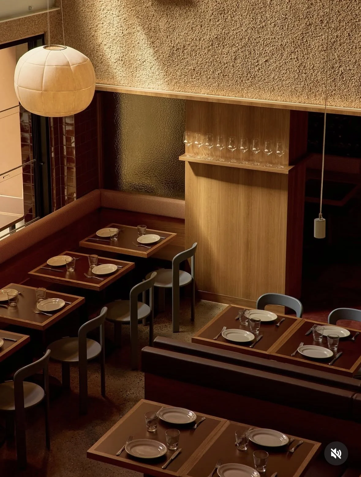 Interior view of a restaurant dining area with wooden tables set with plates, glasses, and silverware, and gray chairs surrounding the tables. There is a large hanging lamp and textured wall surfaces.