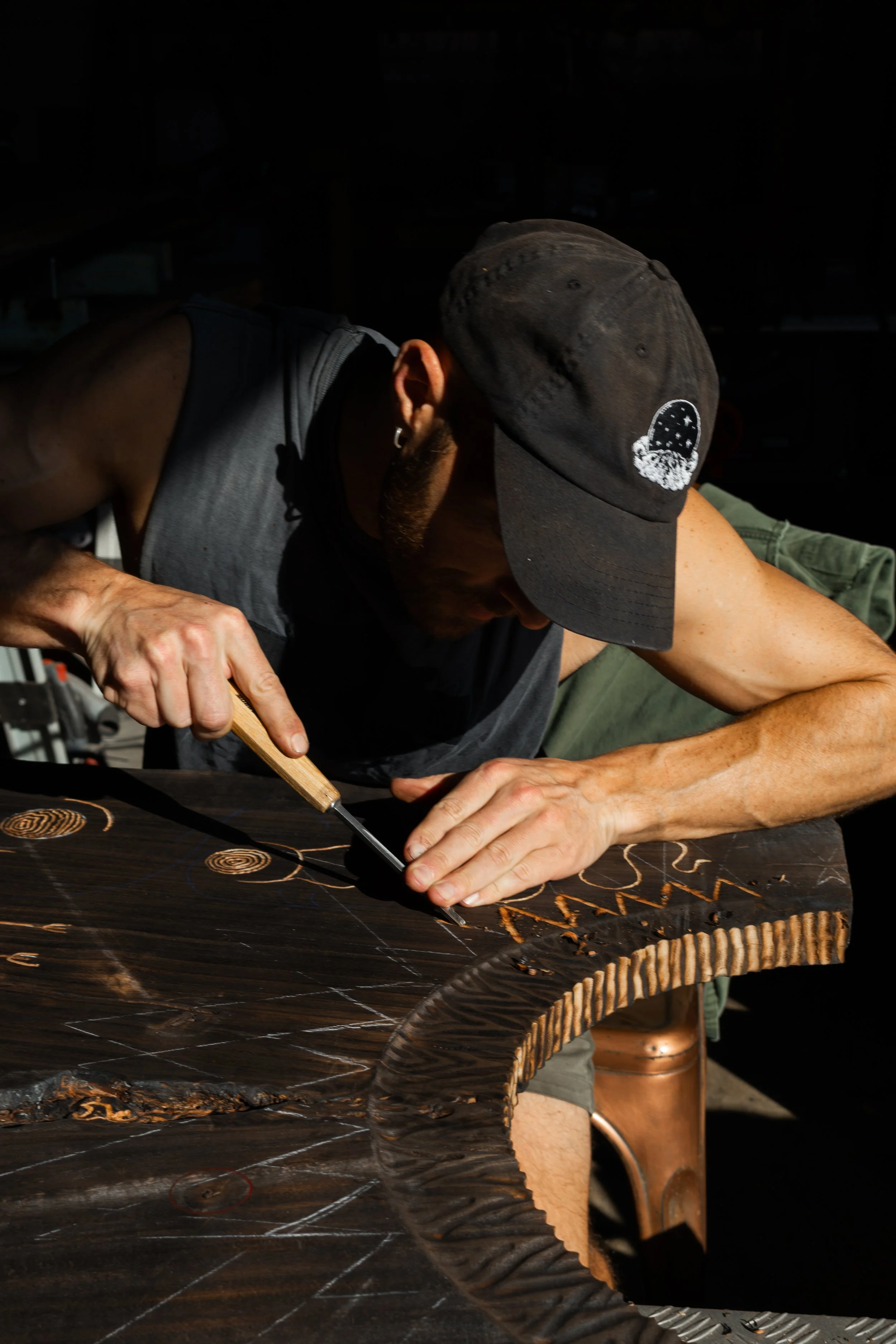 A man wearing a black cap and sleeveless shirt carving designs into a dark piece of wood with a chisel.