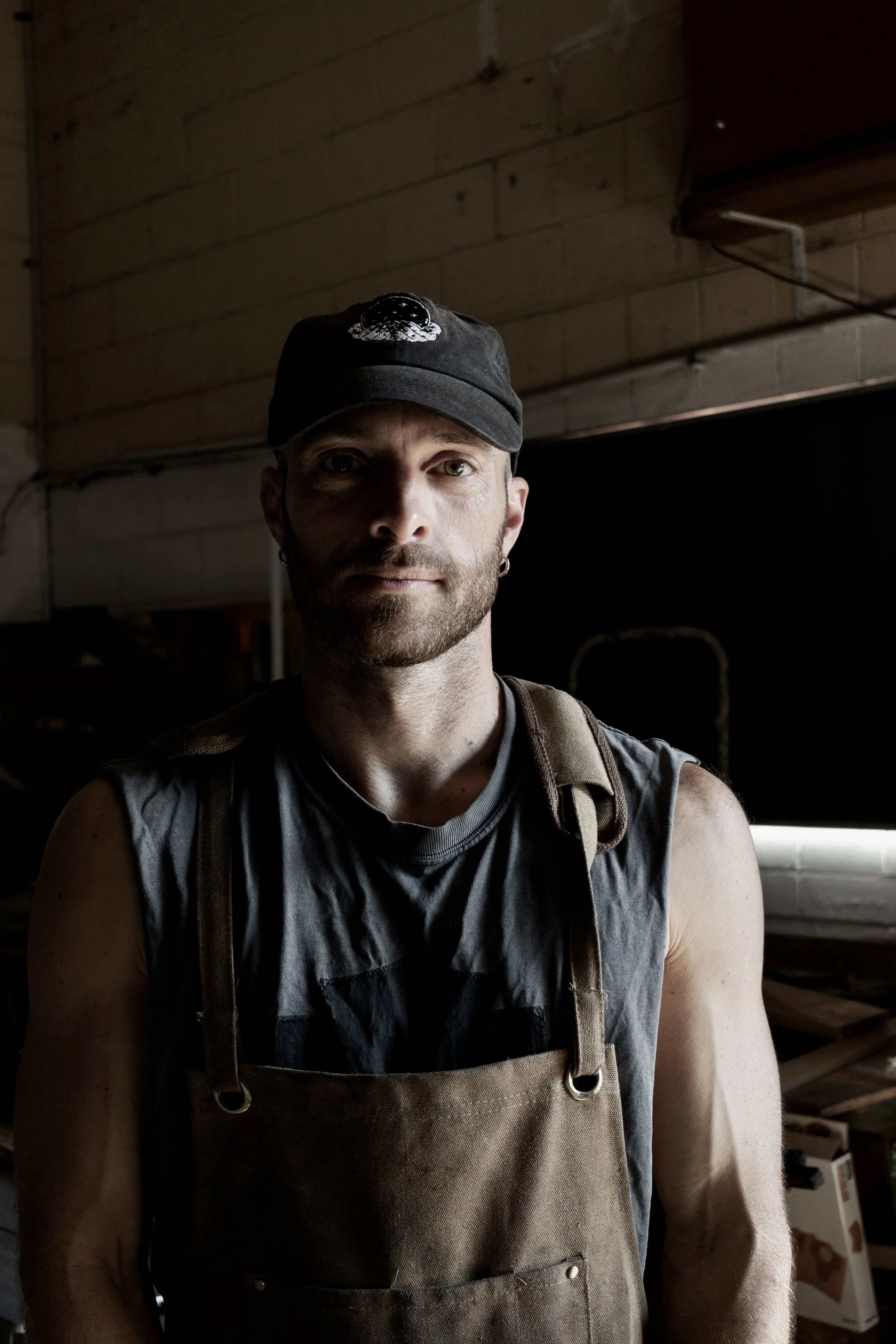 A man wearing a black cap, sleeveless shirt, and a brown apron stands indoors in a workshop or garage, with a serious expression on his face.