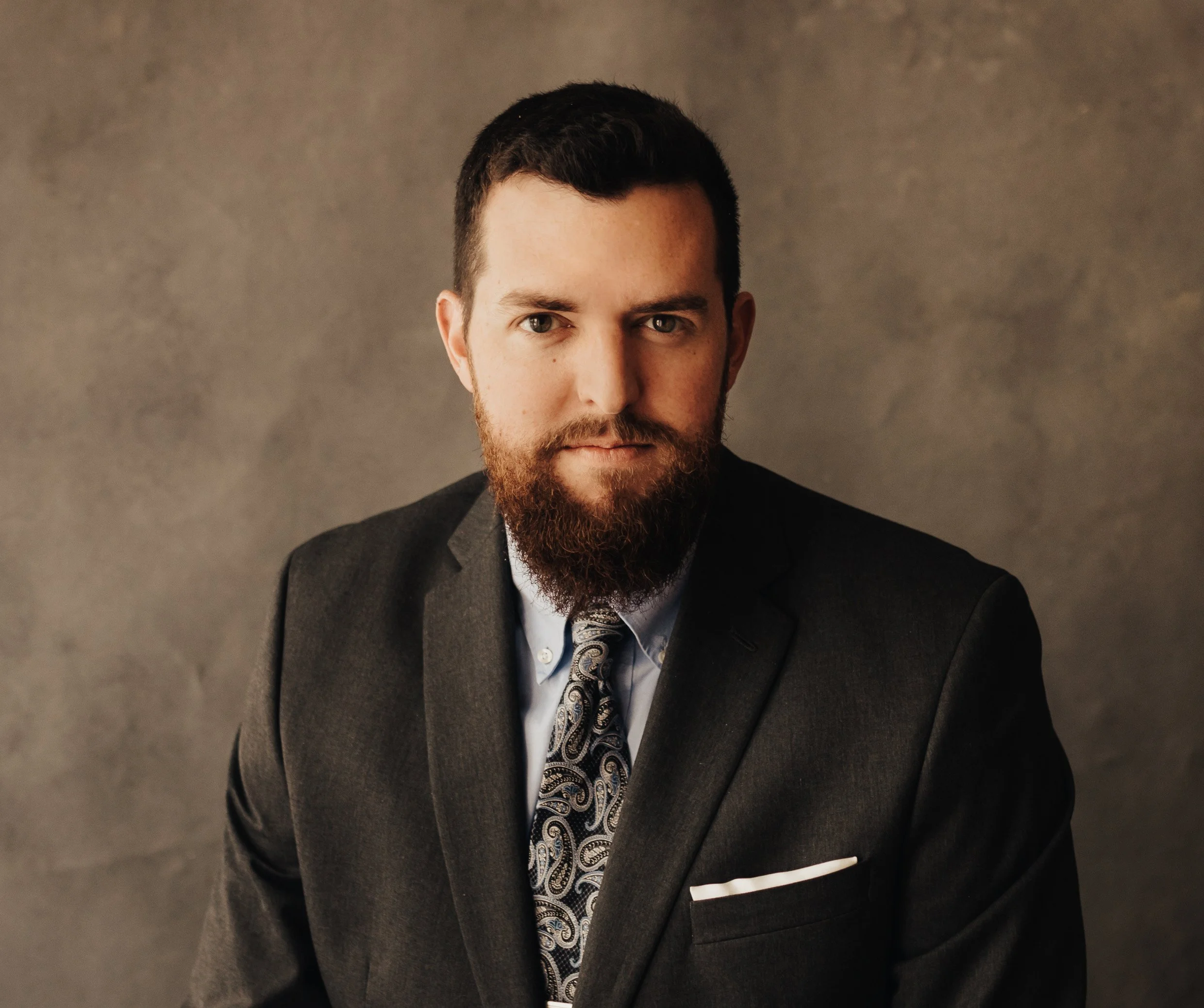 A man in a dark suit with a patterned tie and a light blue dress shirt poses against a neutral background.