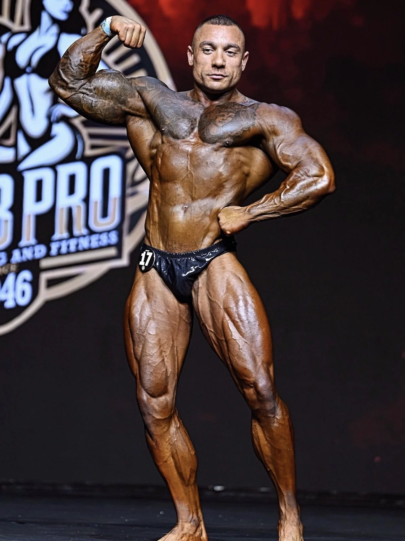 A male bodybuilder in black posing trunks flexes his right bicep on stage during a bodybuilding competition, with a dark background and a logo in the back.