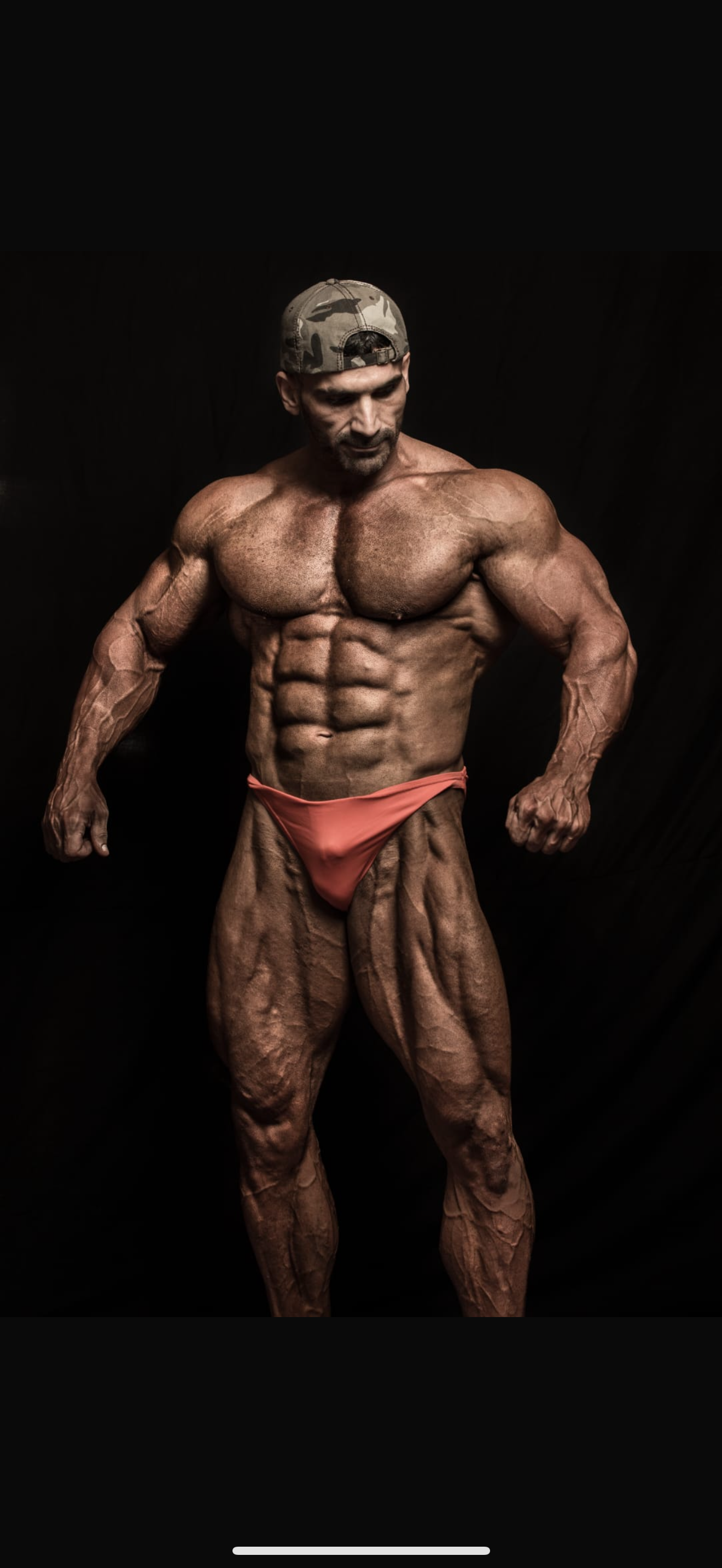 A muscular man wearing a camouflage baseball cap and peach-colored posing trunks, showcasing his defined physique against a black background.