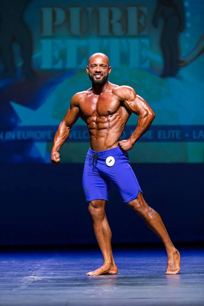 A muscular man with a beard in blue shorts posing on stage at a fitness competition.