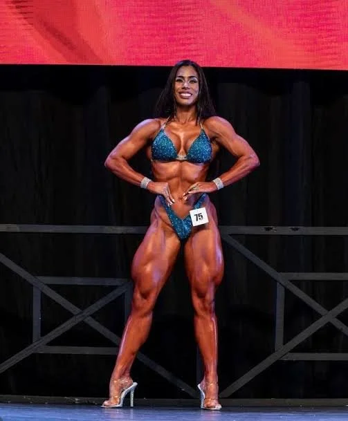 A female bodybuilder in a blue bikini on stage during a competition, flexing her muscles and smiling.