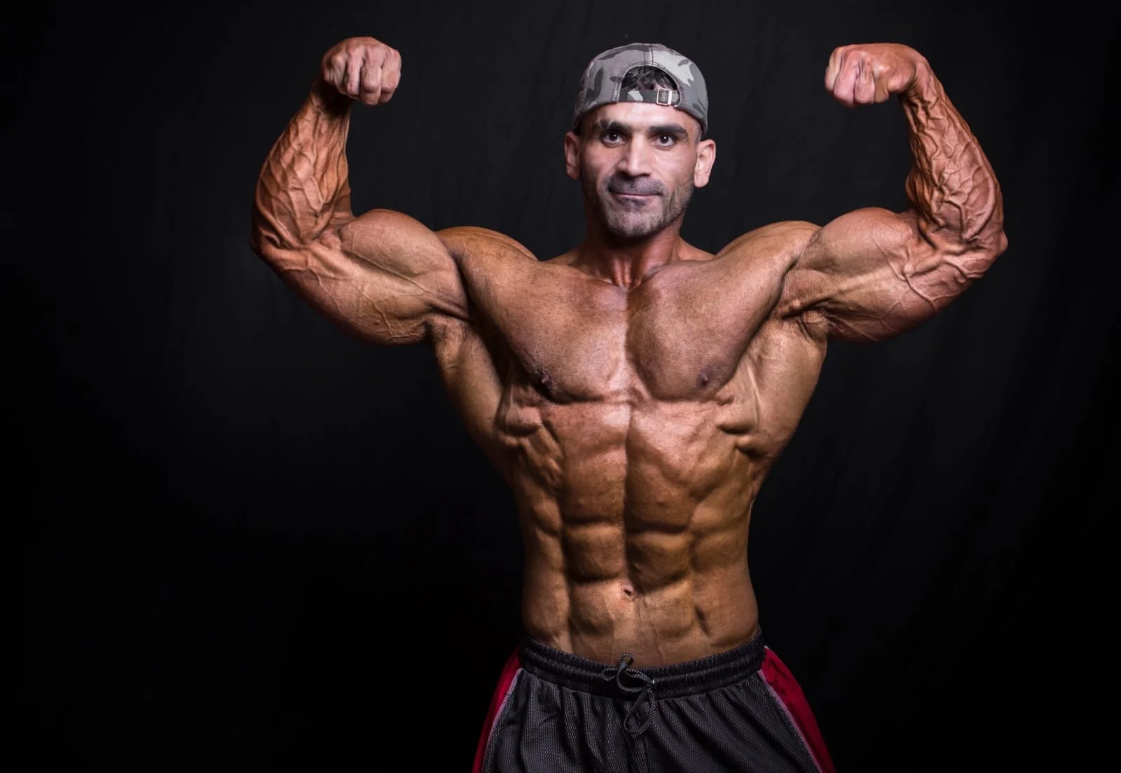 A muscular man wearing a camouflage cap, flexing his biceps against a black background.