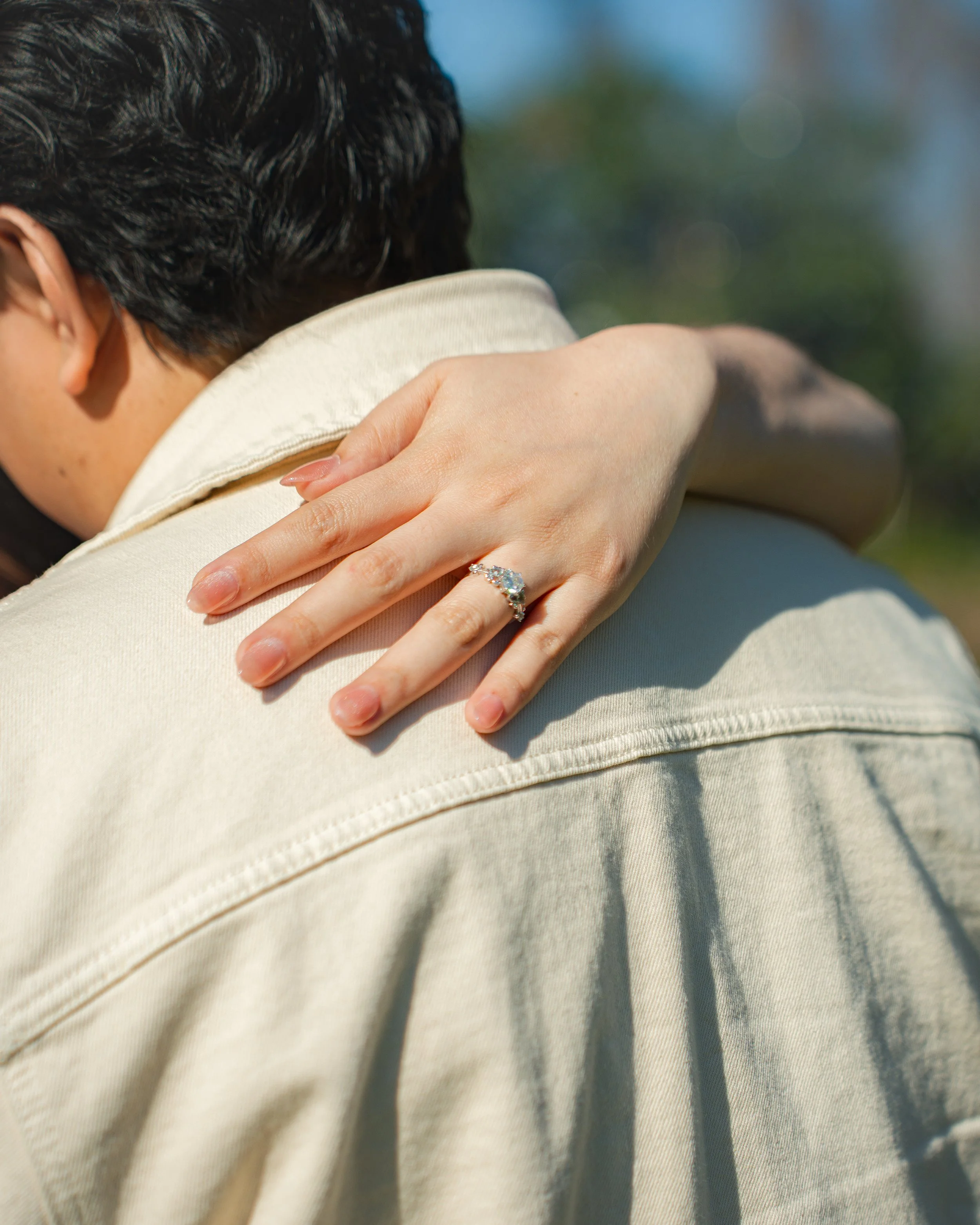 Close-up of a woman’s hand with an engagement ring resting on another person’s shoulder in bright outdoor sunlight.