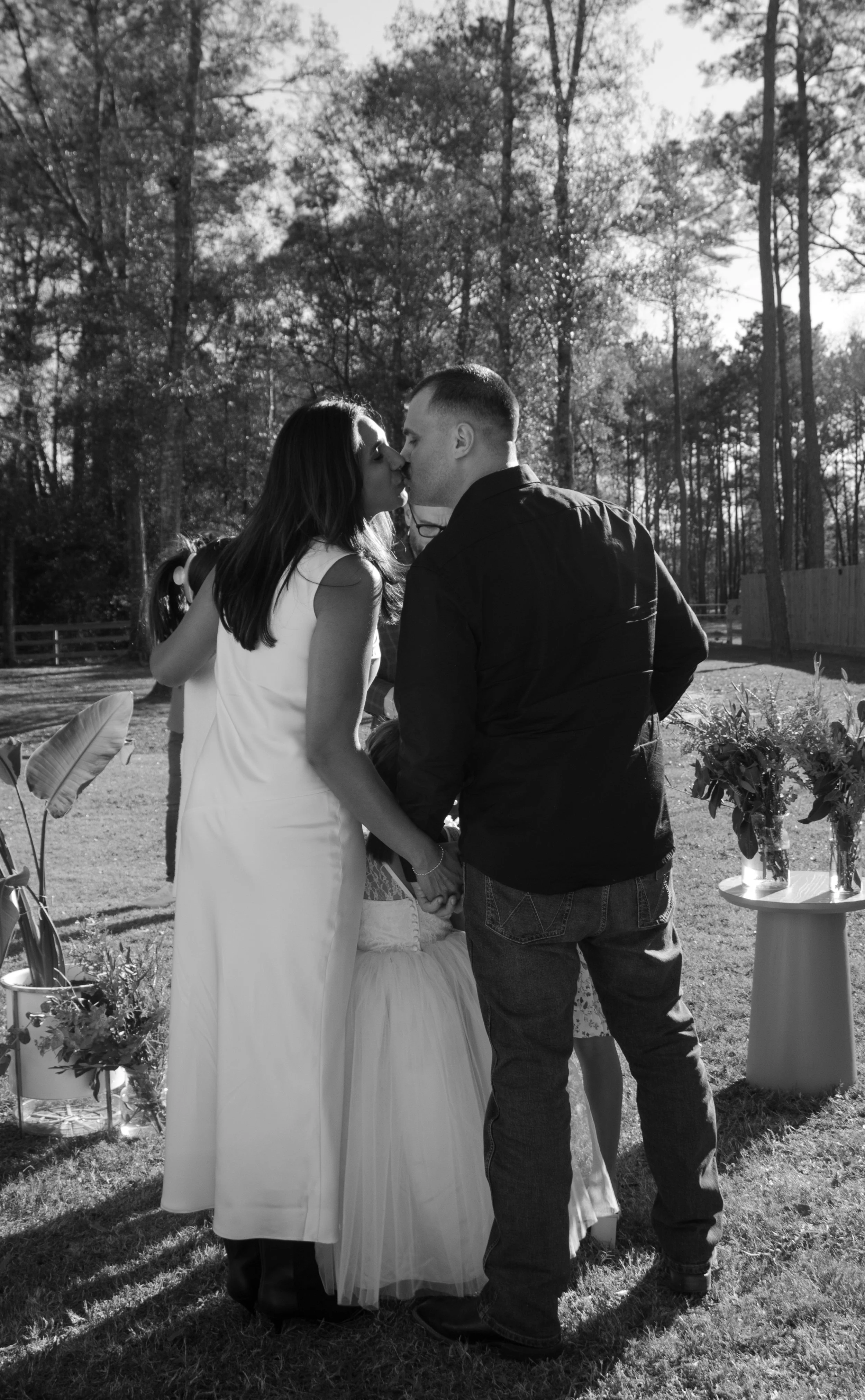 A couple dressed in wedding attire sharing a kiss outdoors surrounded by trees, with a woman in a white wedding dress and a man in a dark shirt and jeans, holding hands during a wedding ceremony.