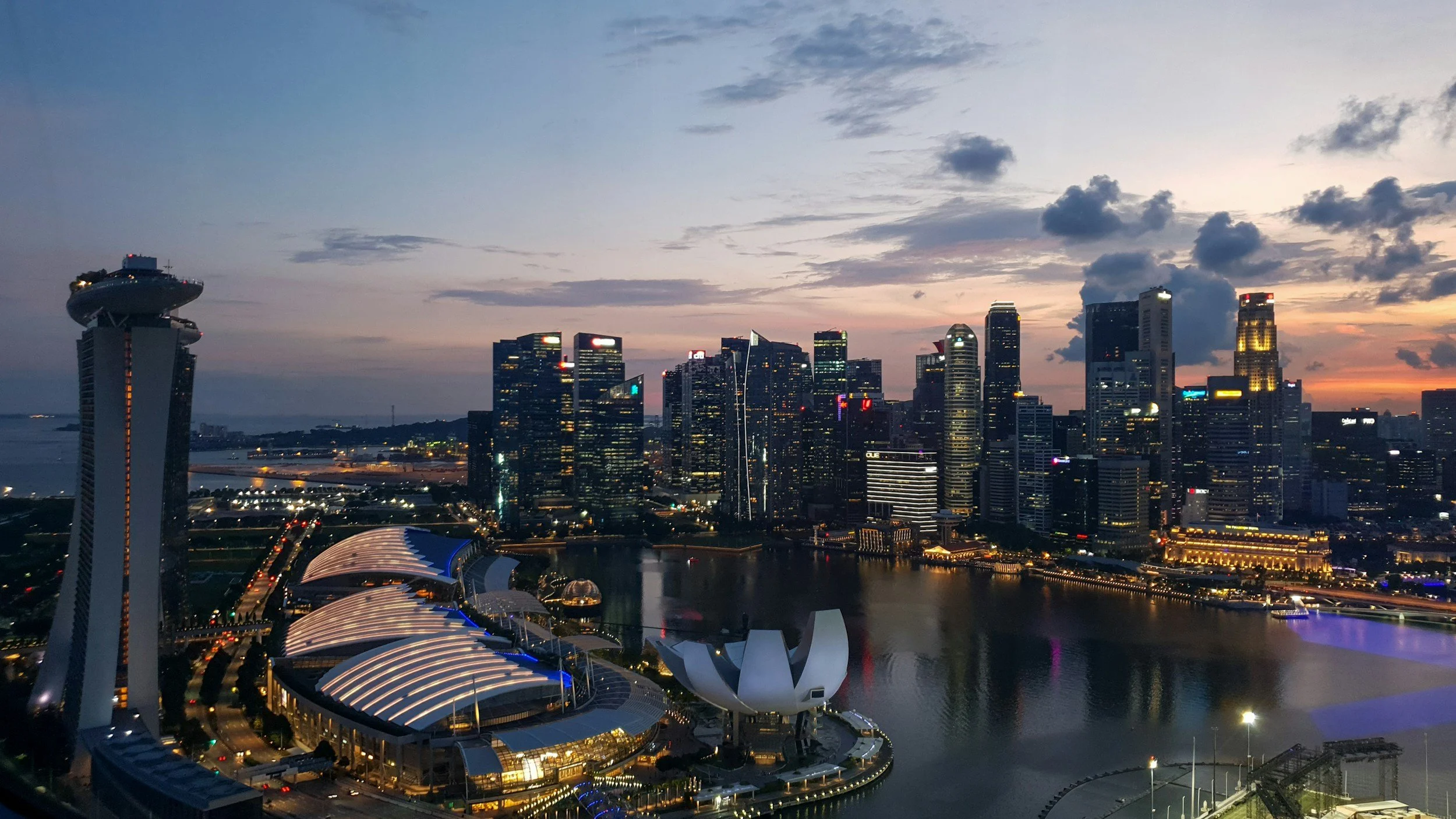 Singapore skyline at dusk with lit buildings, the Marina Bay Sands hotel on the left, the ArtScience Museum in the foreground, and water reflecting the city lights.