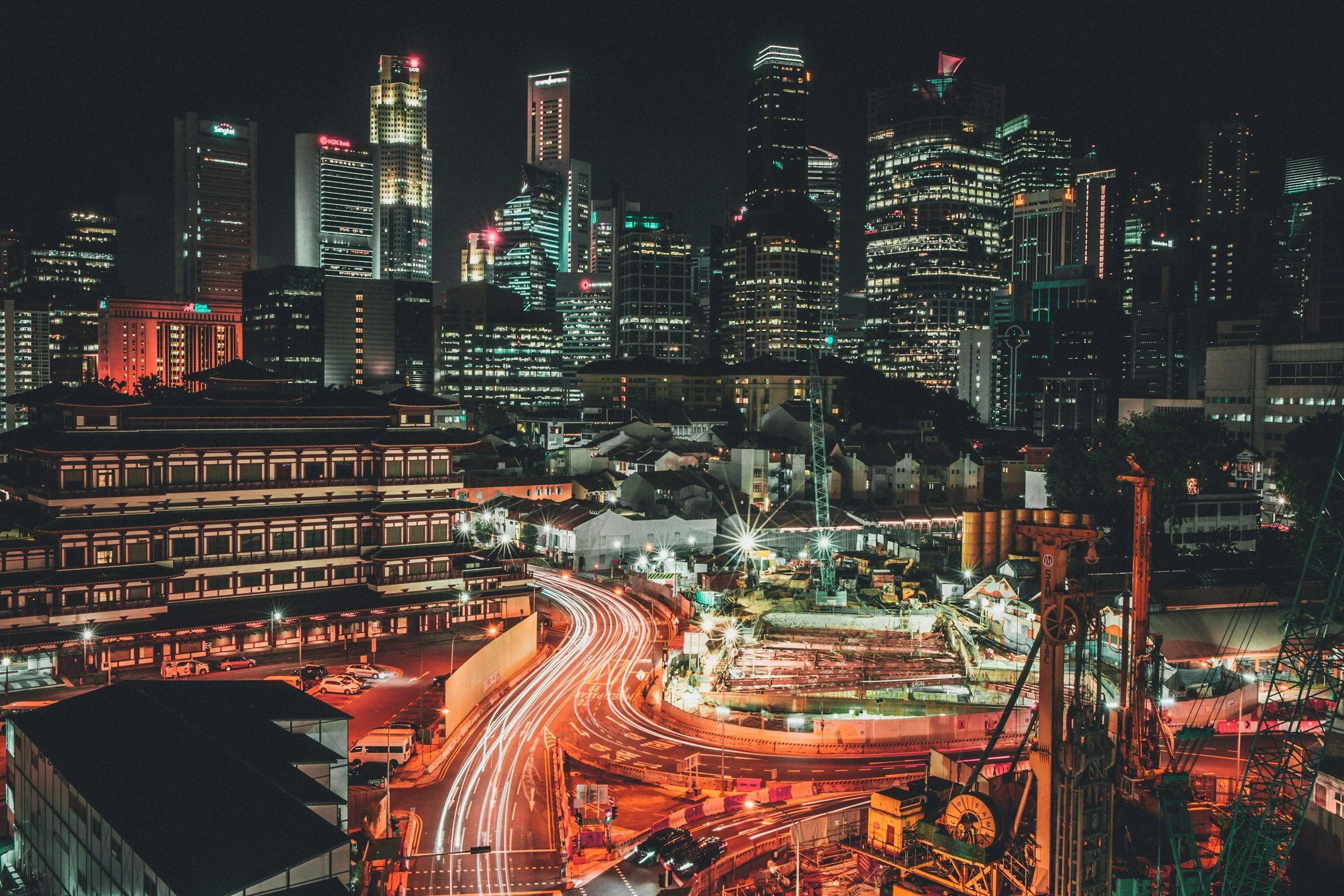 Nighttime cityscape with illuminated skyscrapers, a construction site with equipment, and light trails from moving vehicles.