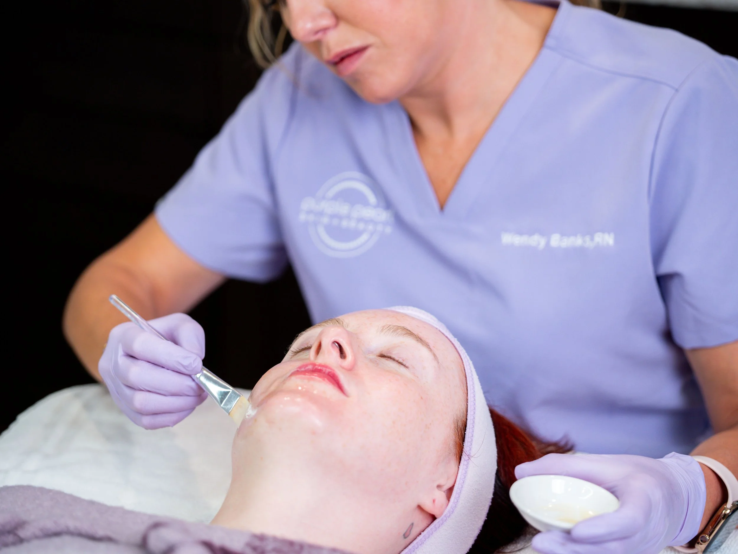 A woman in a light blue medical uniform performing a cosmetic facial treatment on a client lying down with closed eyes. The woman is using a tool on the client's face in a clinical setting.