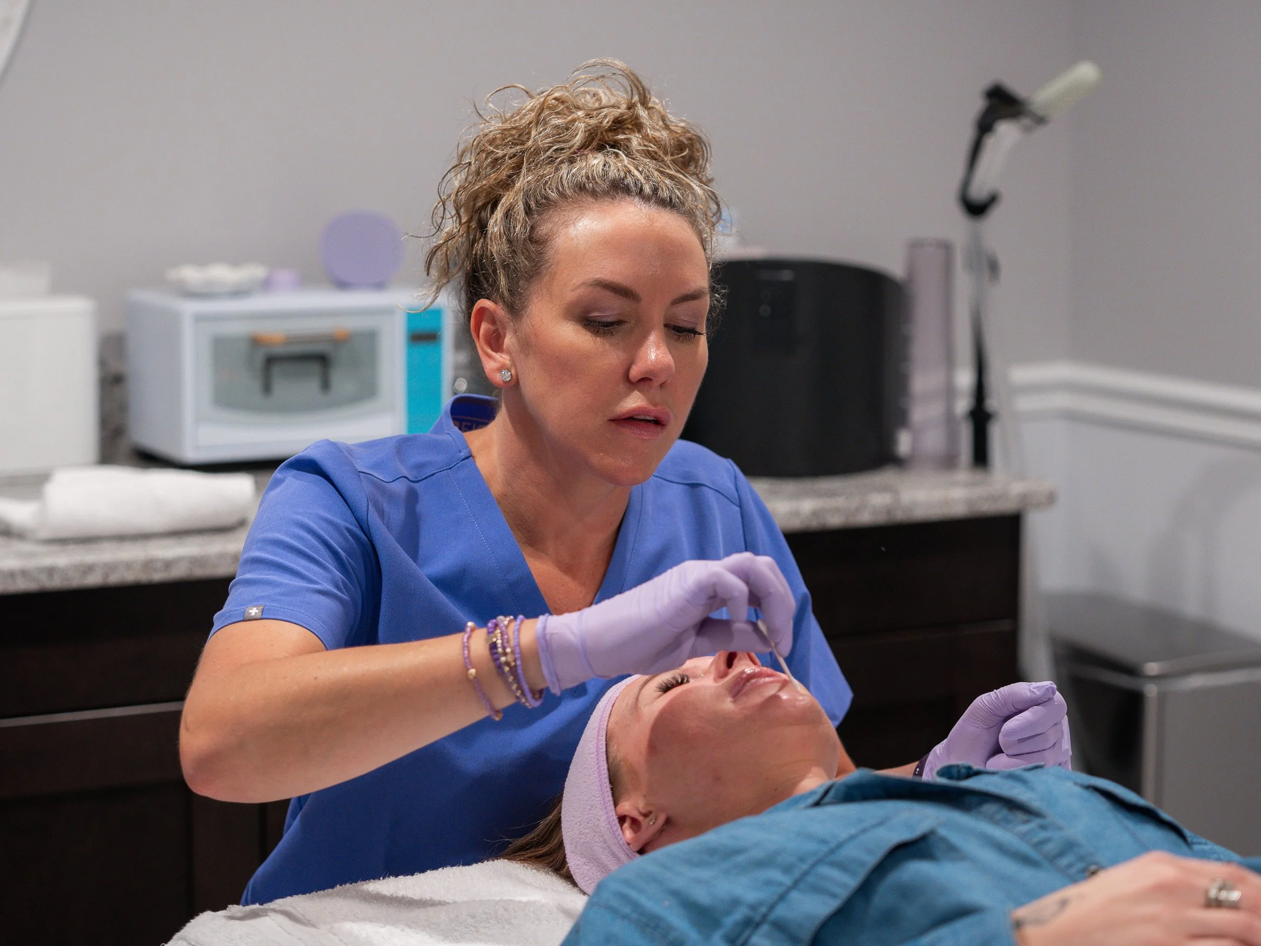 A female medical professional in blue scrubs giving a procedure or treatment to a person lying down, in a clinical setting.