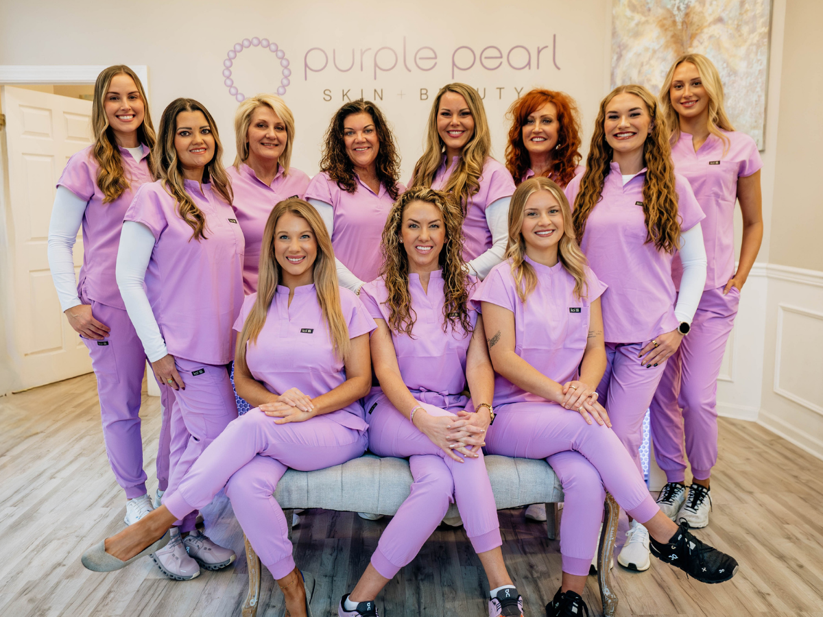 Group photo of ten women wearing lavender scrubs, sitting and standing in front of a sign that reads 'Purple Pearl Skin + Beauty' inside a spa or clinic.