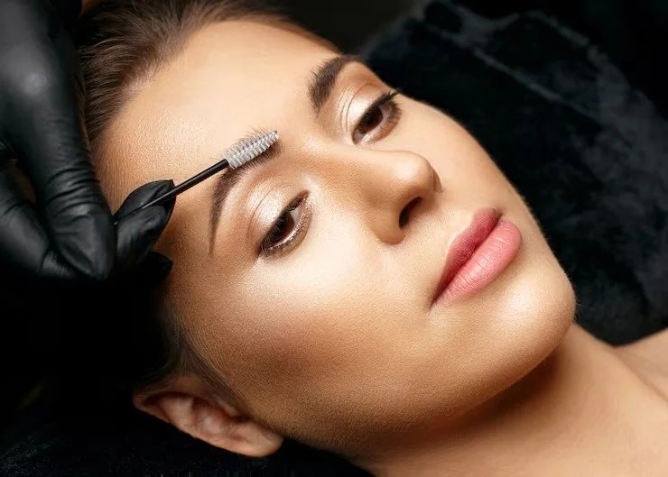 Close-up of a woman receiving eyelash or eyebrow grooming treatment, with a stylist applying product with a small brush on her eyebrow.