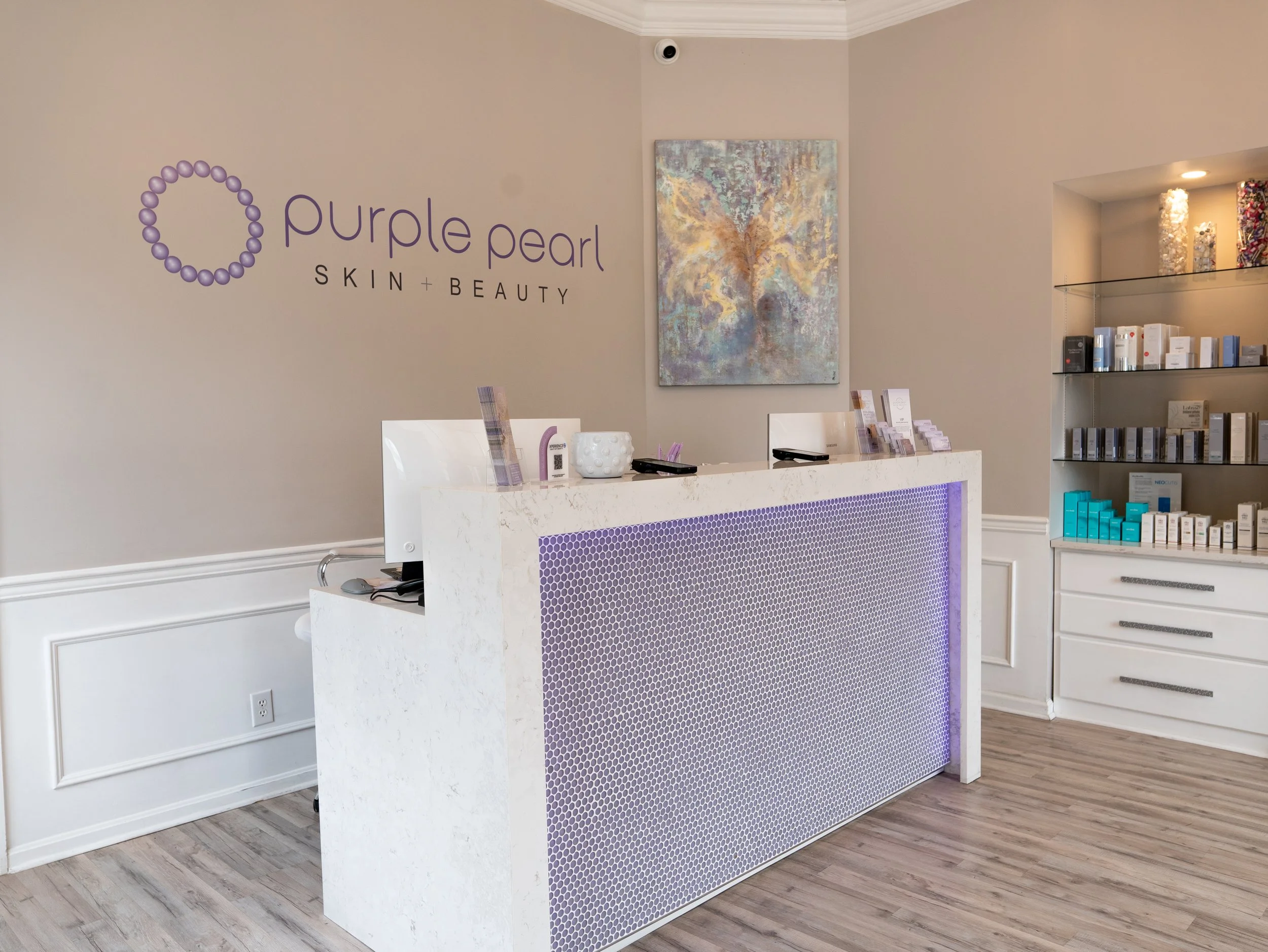 Interior view of a skincare and beauty clinic reception area with a white marble counter, purple accents, a wall sign for 'Purple Pearl Skin + Beauty,' a colorful abstract painting, and shelves with skincare products.
