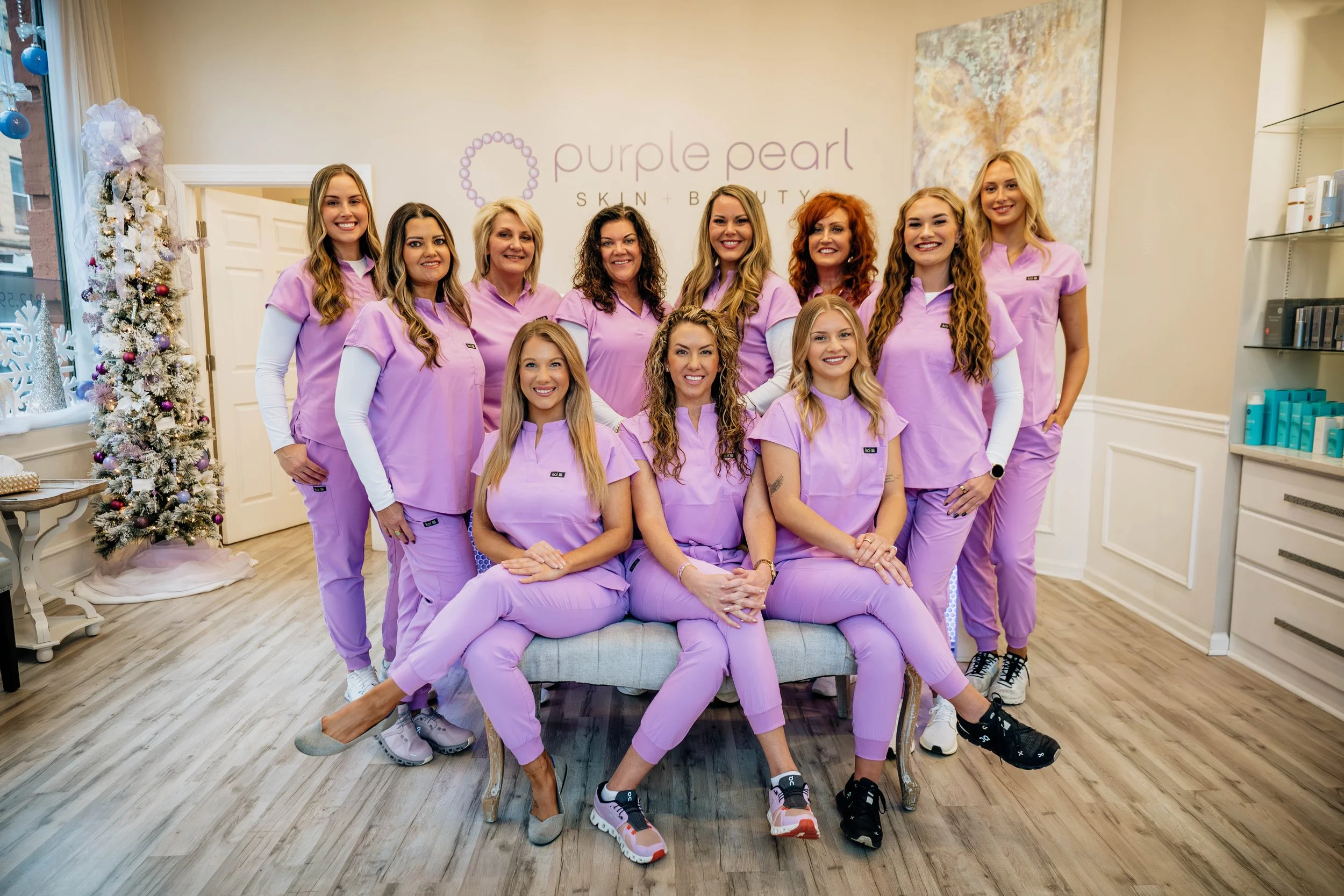 Group of women wearing matching purple scrubs posing for a photo inside a beauty or skincare clinic, with a decorated Christmas tree in the background.