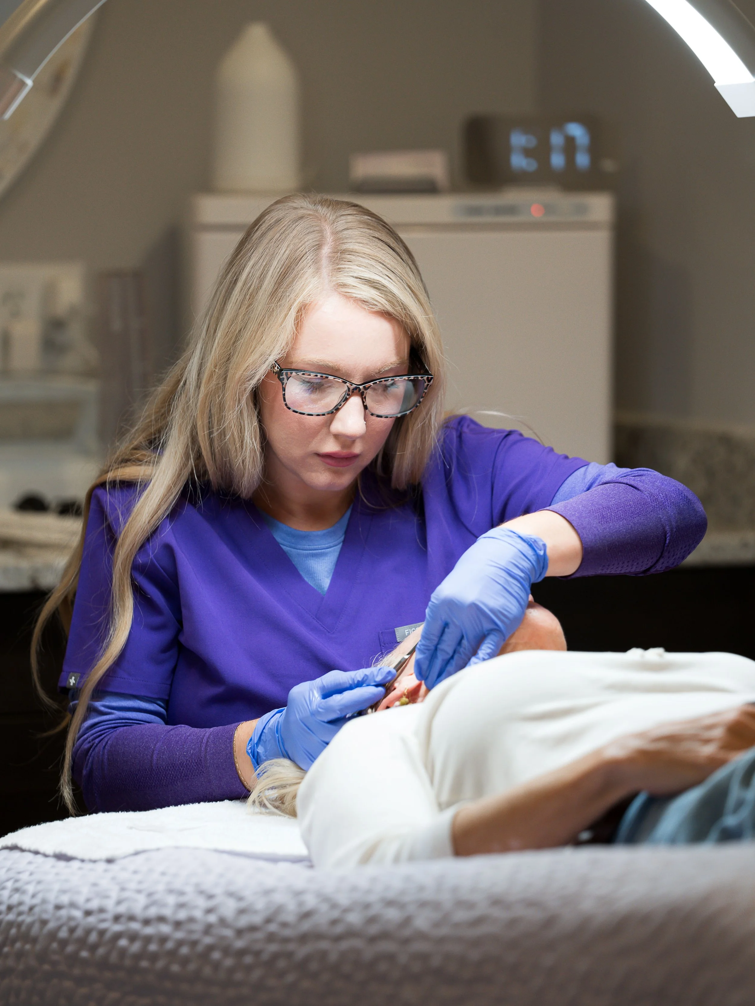 A female medical professional wearing glasses and purple scrubs performing a medical procedure on a patient in a clinical setting.