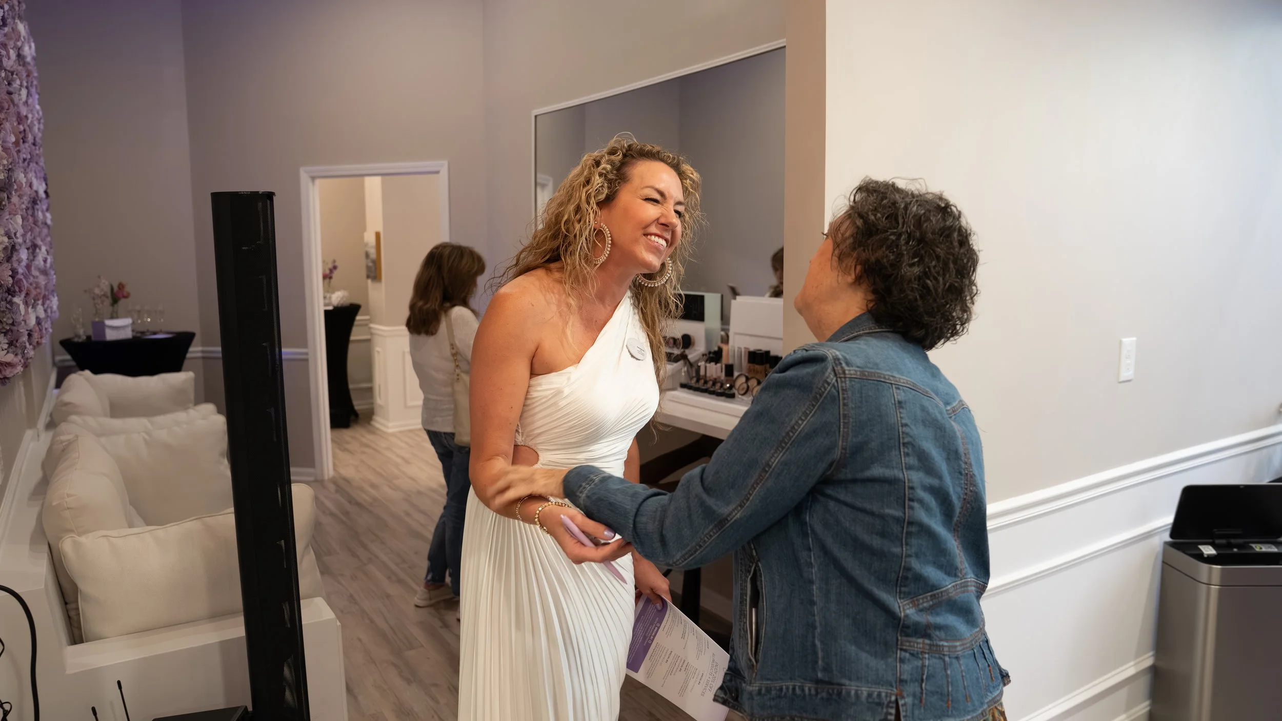 A woman in a white dress smiling as she is greeted by another woman in a denim jacket at an indoor event.
